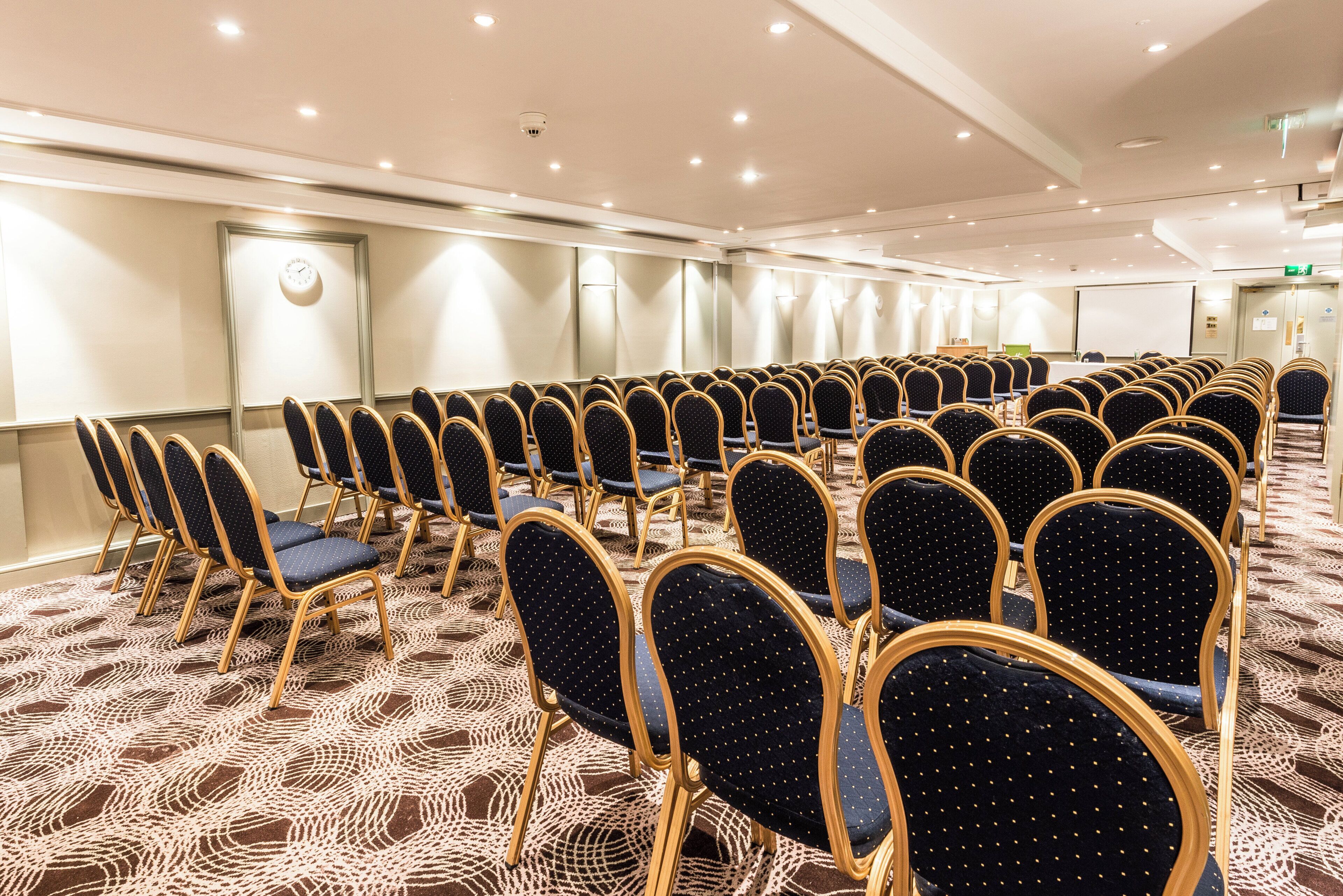 A conference room with rows of chairs, a clock, and a projector screen.