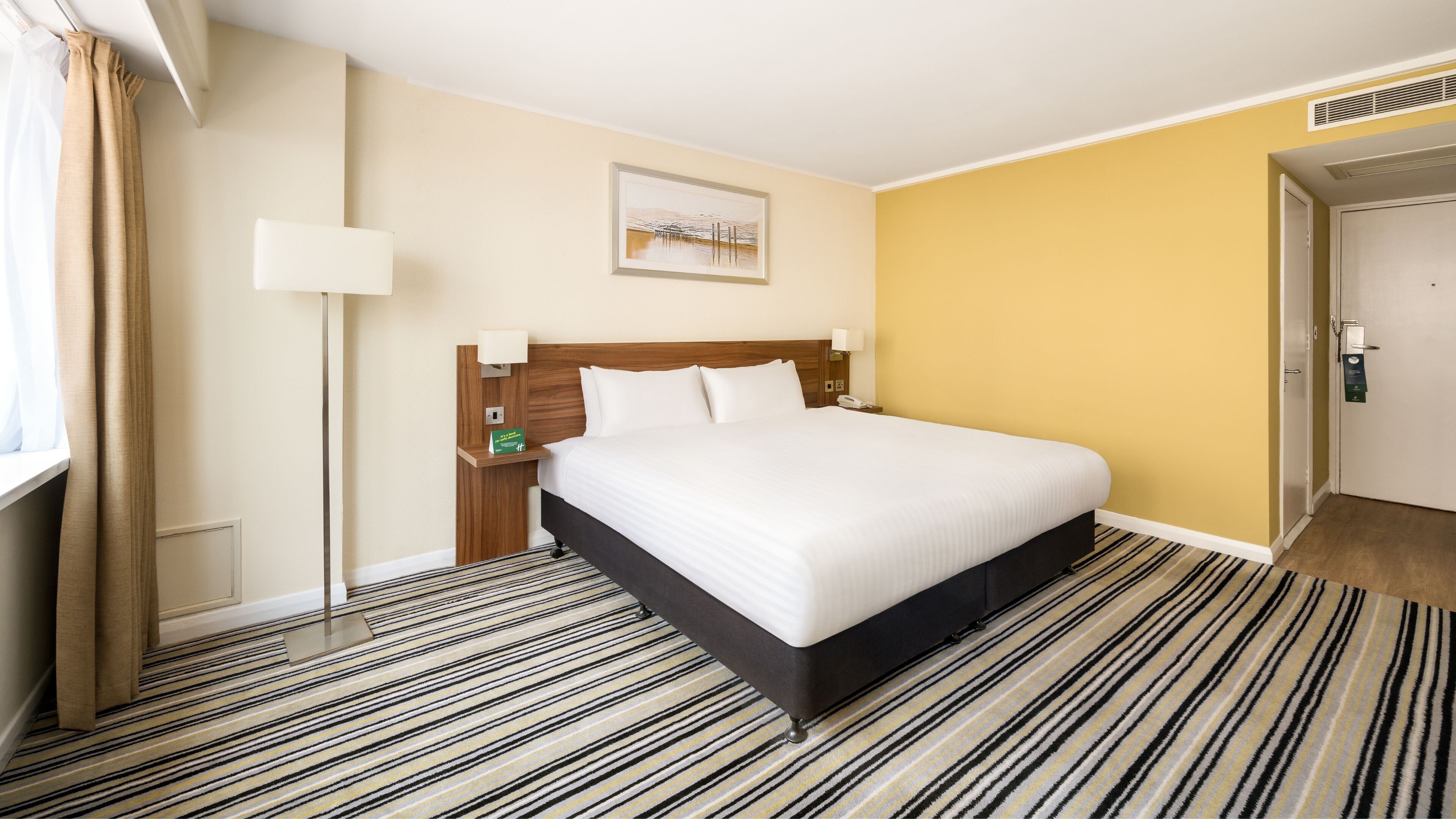 A hotel room with a large bed, a bedside table with a lamp, a painting on the wall, and a striped carpet.