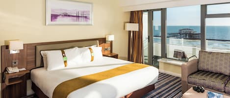 A hotel room with a large bed, a view of the sea, a desk with a lamp, and a chair.