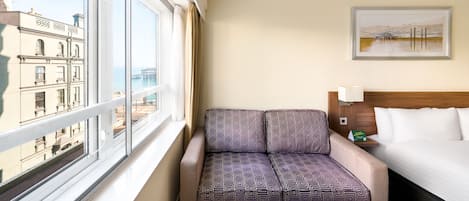 A hotel room with a bed, a window with a view of buildings, a sofa with patterned cushions, and a framed picture on the wall.