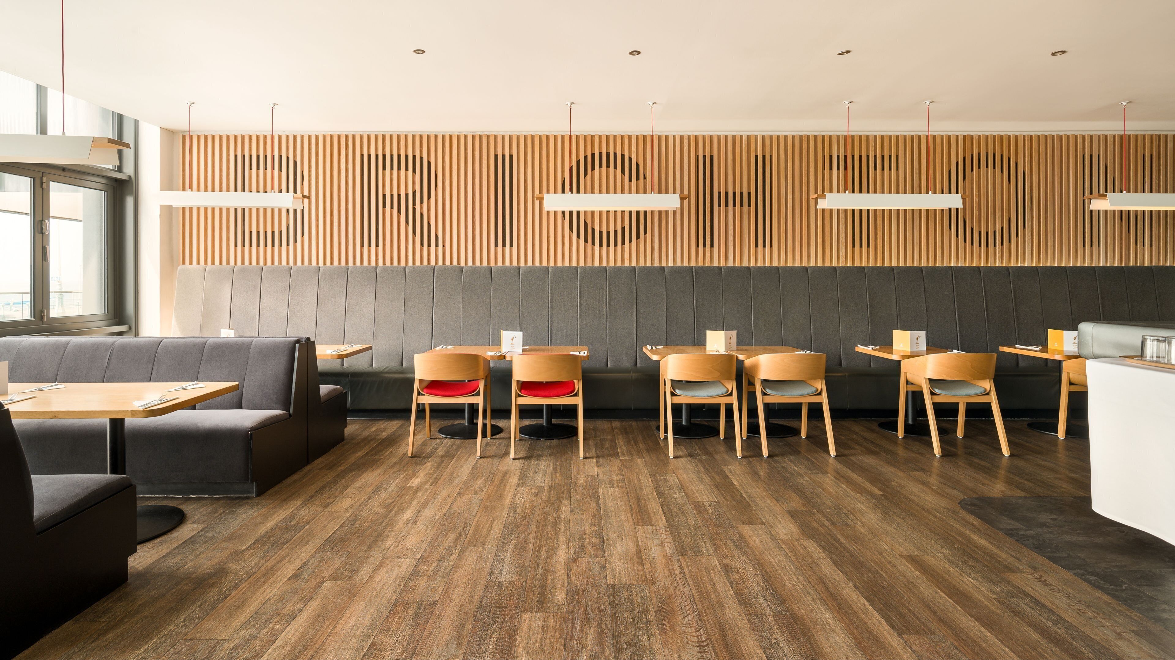 Un intérieur de restaurant moderne avec des murs lambrissés en bois, des sièges rembourrés de couleur grise et des tables en bois.