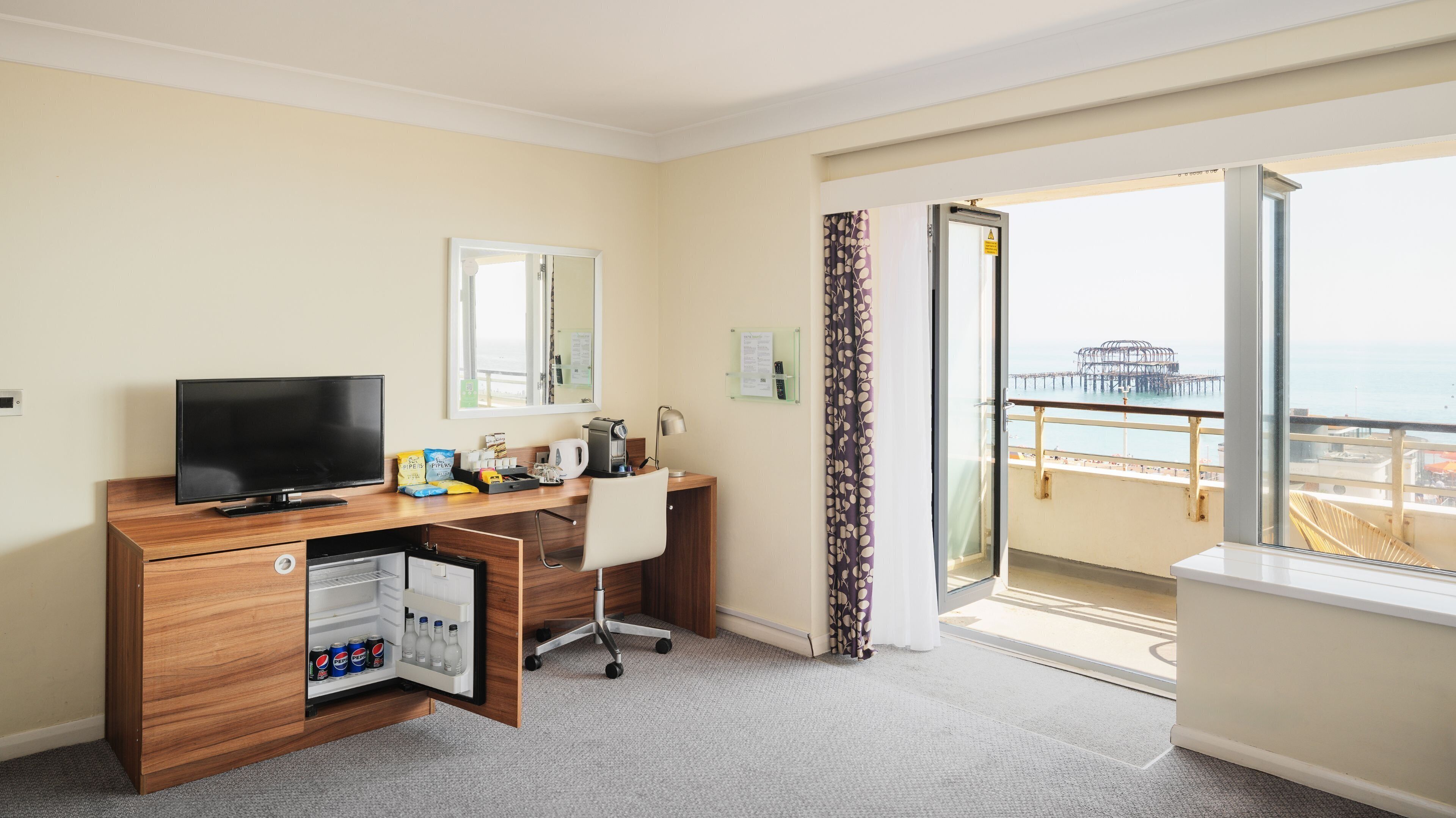 Ein Hotelzimmer mit Holzschreibtisch, Fernseher, einer Minibar mit Getränken, einem Sessel, einem Fenster mit Blick auf einen Steg und einem Vorhang.