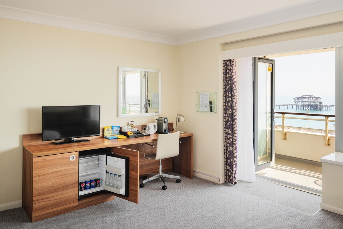 Une chambre d’hôtel équipée d’un bureau en bois, d’une télévision, d’un mini-réfrigérateur contenant des boissons, d’une chaise, d’une fenêtre donnant sur une jetée et d’un rideau.