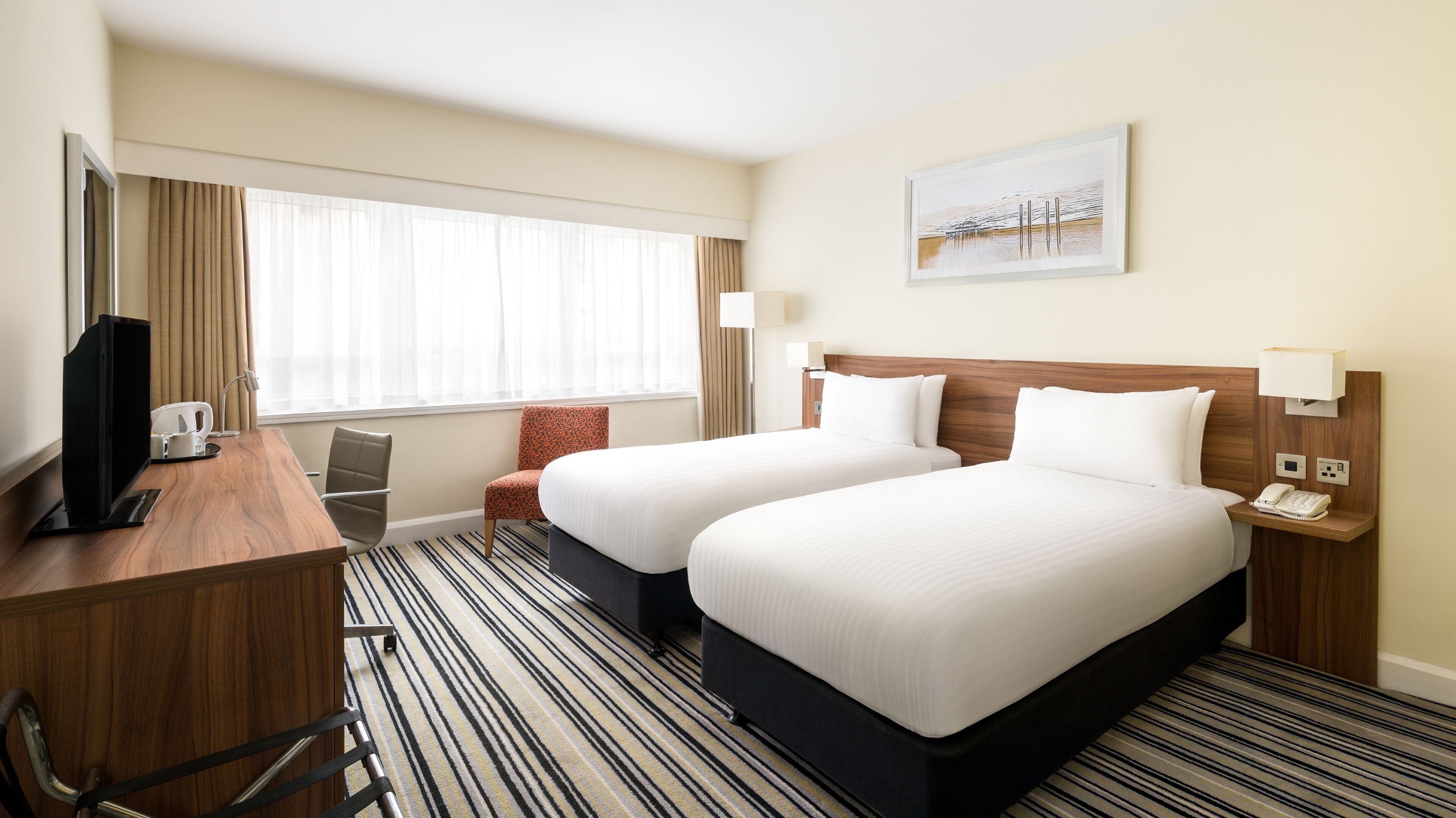 A hotel room with two beds, a desk, a chair, a TV, and a painting on the wall.