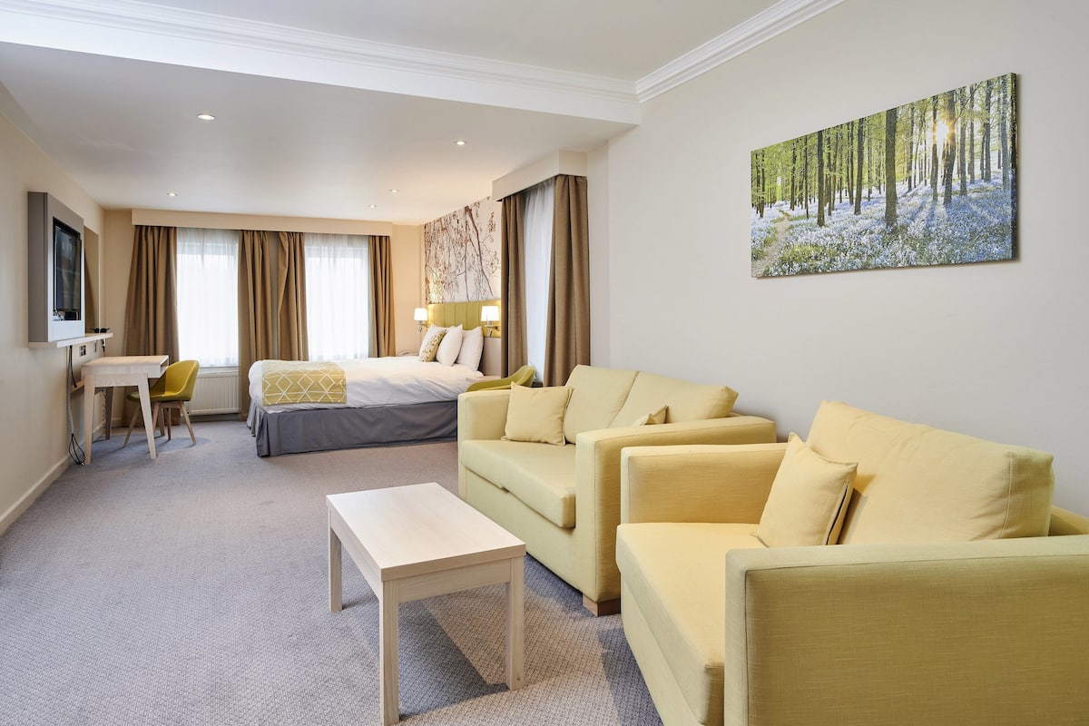 A hotel room with a bed, two yellow sofas, a small table, a desk with a chair, and a painting of a forest on the wall.