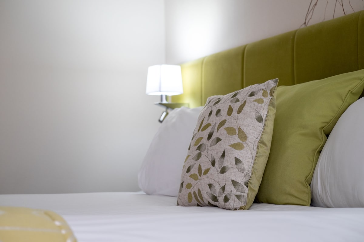 A neatly made bed with white and green pillows, a bedside lamp, and a wall with a bare branch.