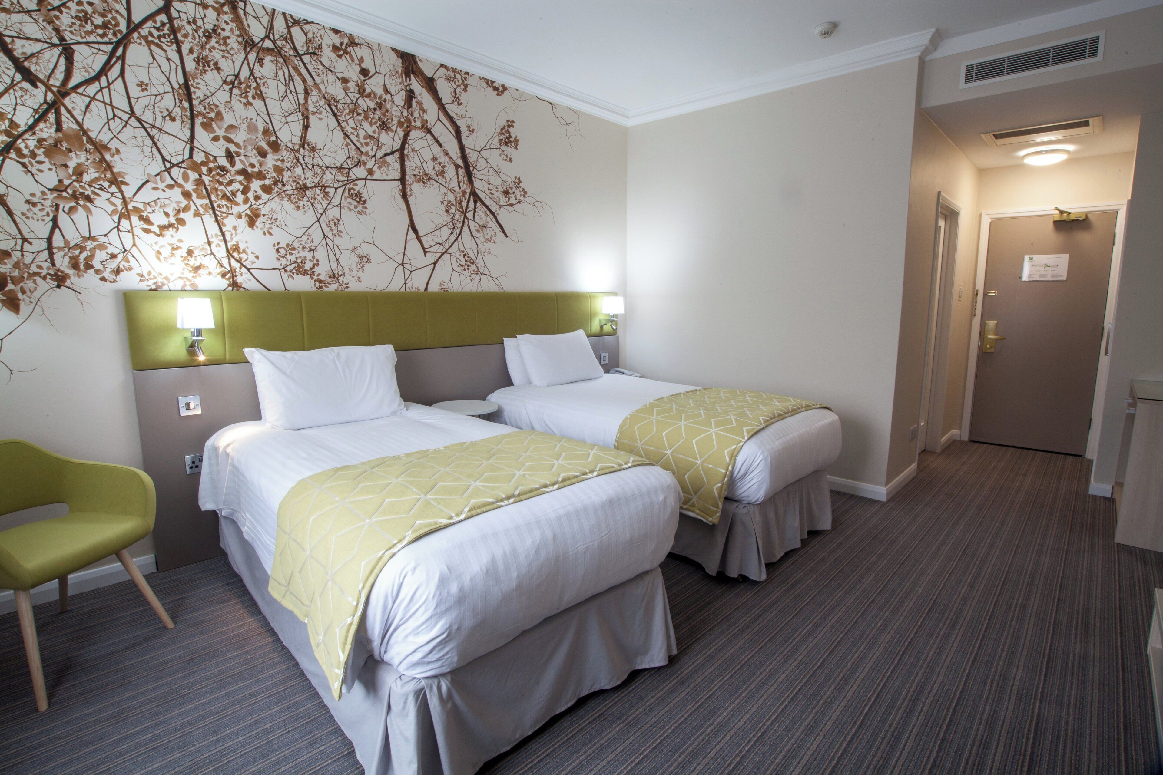 Habitación de hotel con dos camas, una silla verde y un muro con diseño de hojas.