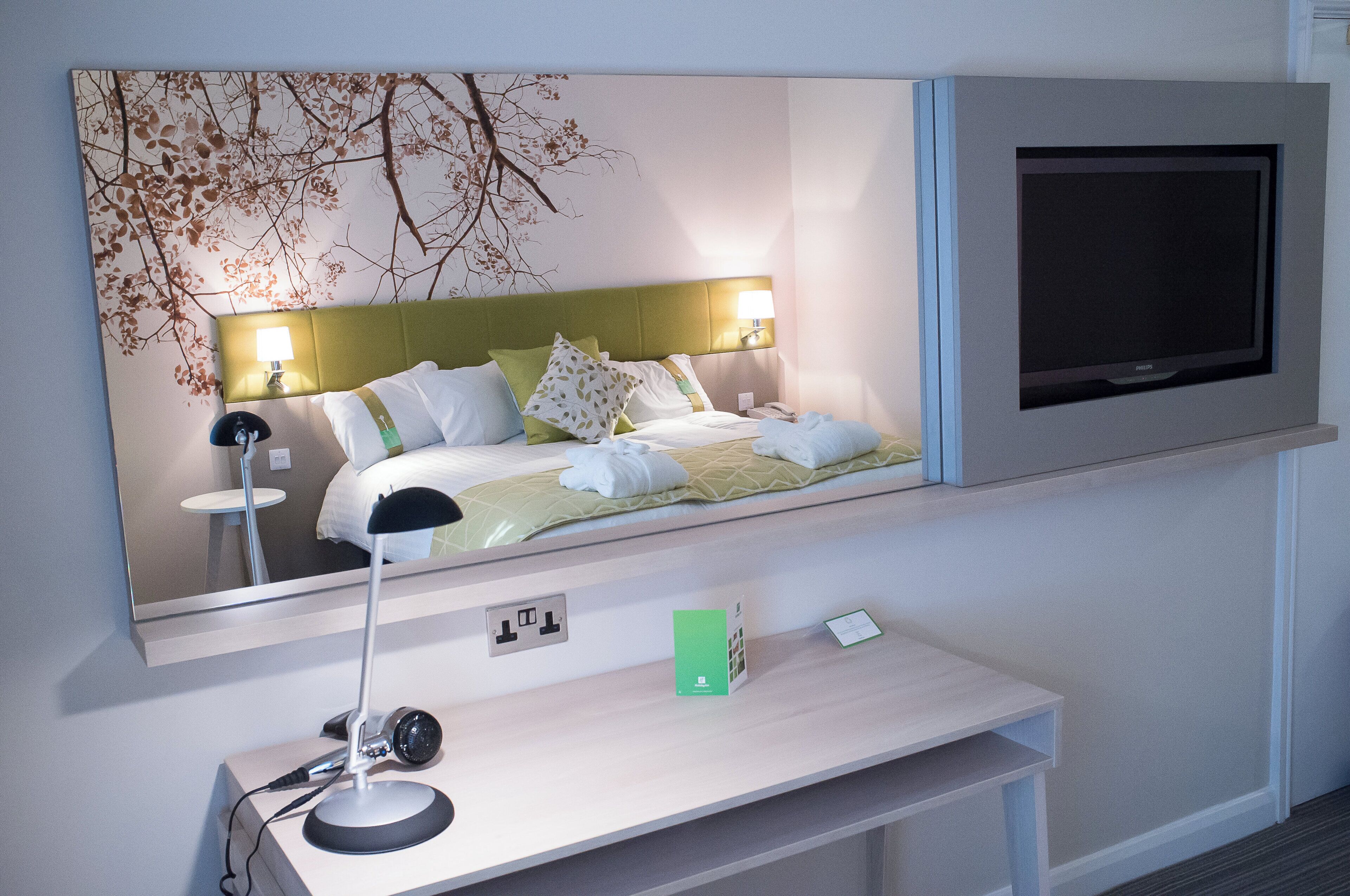 A hotel room with a bed, a bedside lamp, a television mounted on the wall, and a desk with a lamp.
