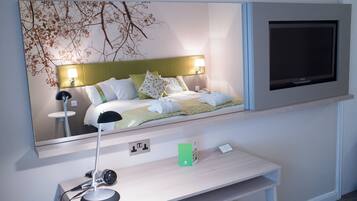 A hotel room with a bed, a bedside lamp, a television mounted on the wall, and a desk with a lamp.