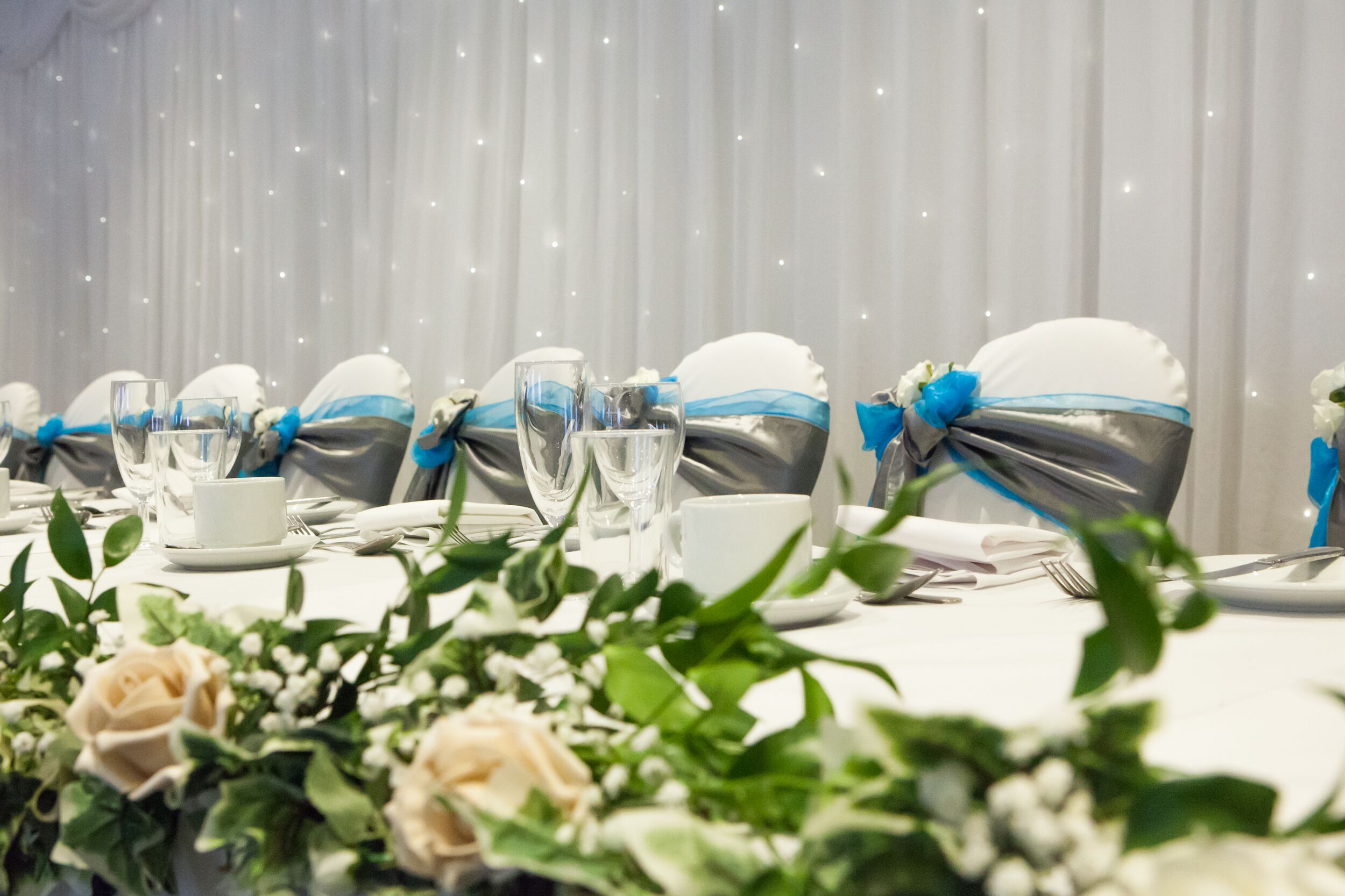Una mesa dispuesta para un evento formal, con manteles blancos, servilletas de plata atadas con cintas azules y cristalería transparente.