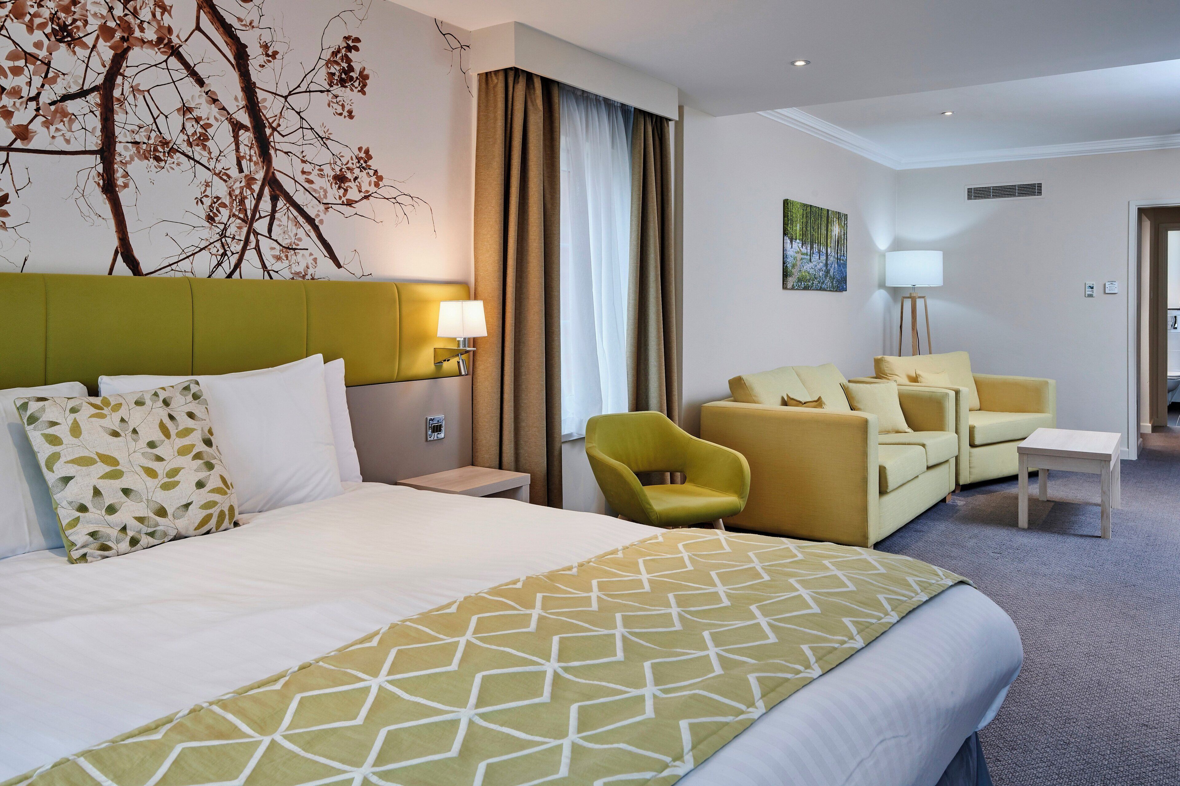 Quarto de hotel com uma cama, duas poltronas, um sofá, uma mesa de cabeceira, um abajur e um quadro na parede.