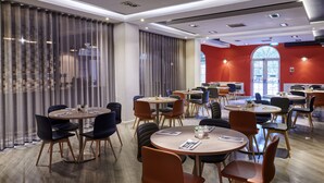 A modern dining area with round tables and chairs, large windows with curtains, and a red accent wall.