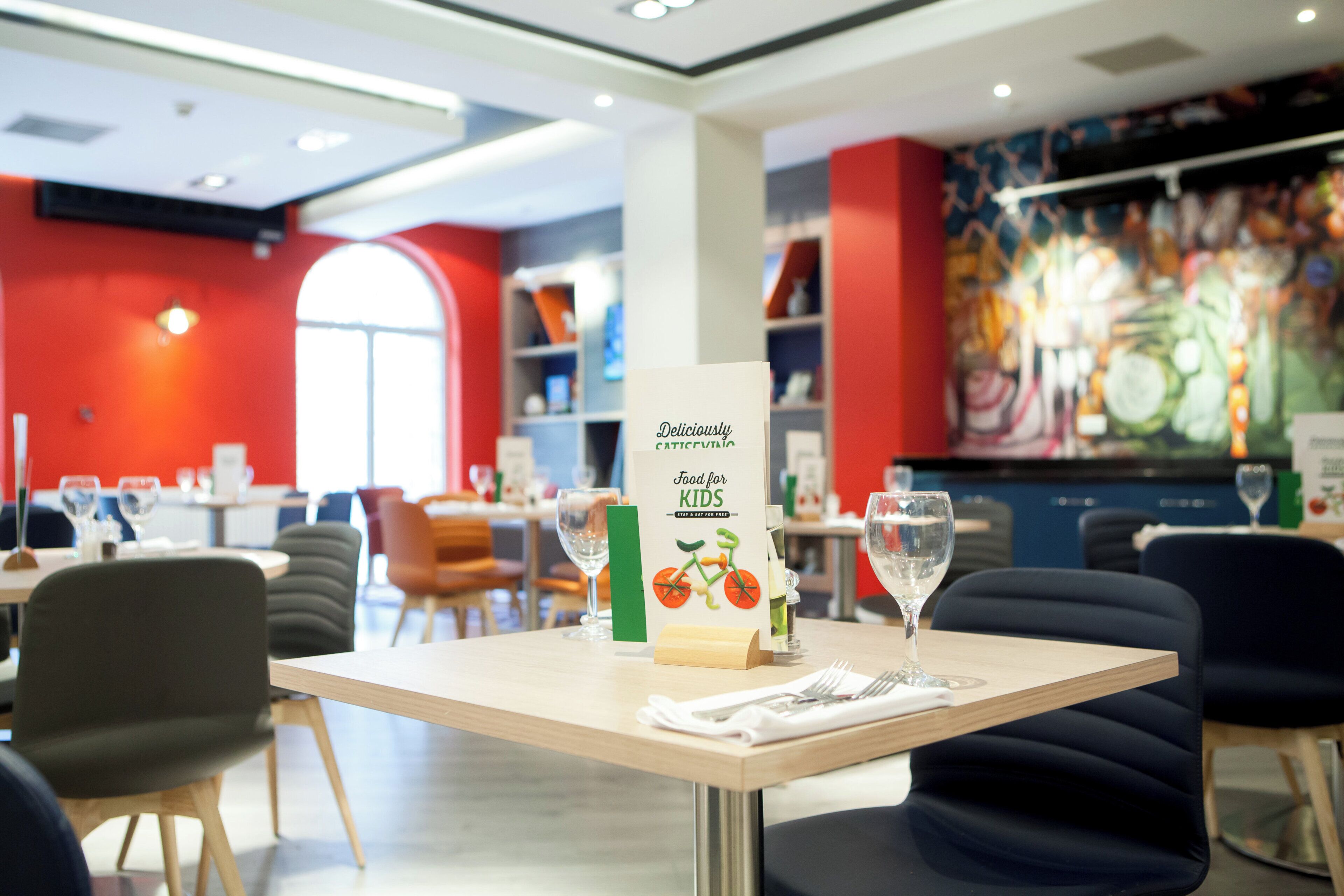 A restaurant interior with a menu stand, dining tables, chairs, and a colorful mural on the wall.