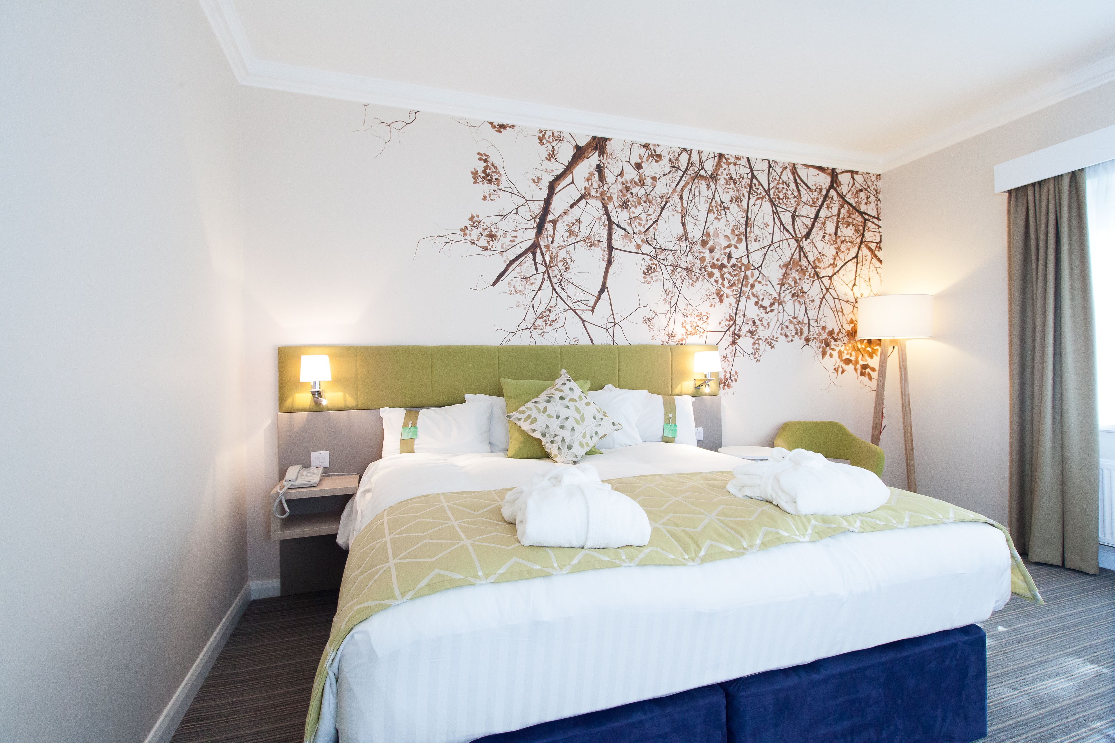 A hotel room with a large bed, a green headboard, a wall-mounted artwork, two bedside lamps, a nightstand, and a telephone.