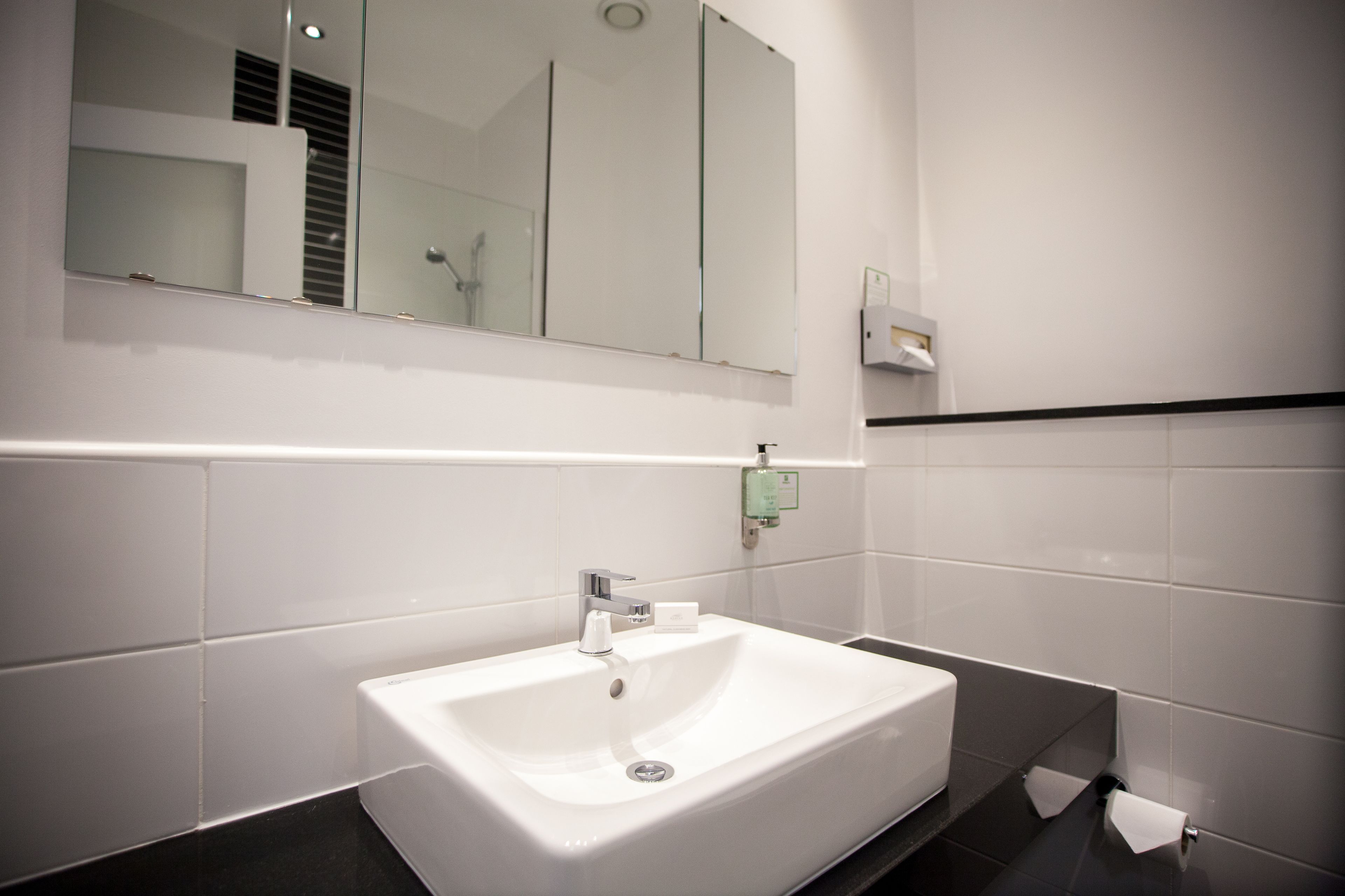 A modern bathroom with a white sink, a mirror above it, and a towel dispenser on the wall.