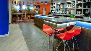A modern bar area with a long counter, red bar stools, and a variety of bottles and glasses on shelves. - Holiday Inn Corby Kettering Hotel & Spa (Corby)
