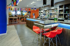 A modern bar area with a long counter, red bar stools, and a variety of bottles and glasses on shelves. - Holiday Inn Corby Kettering Hotel & Spa (Corby)