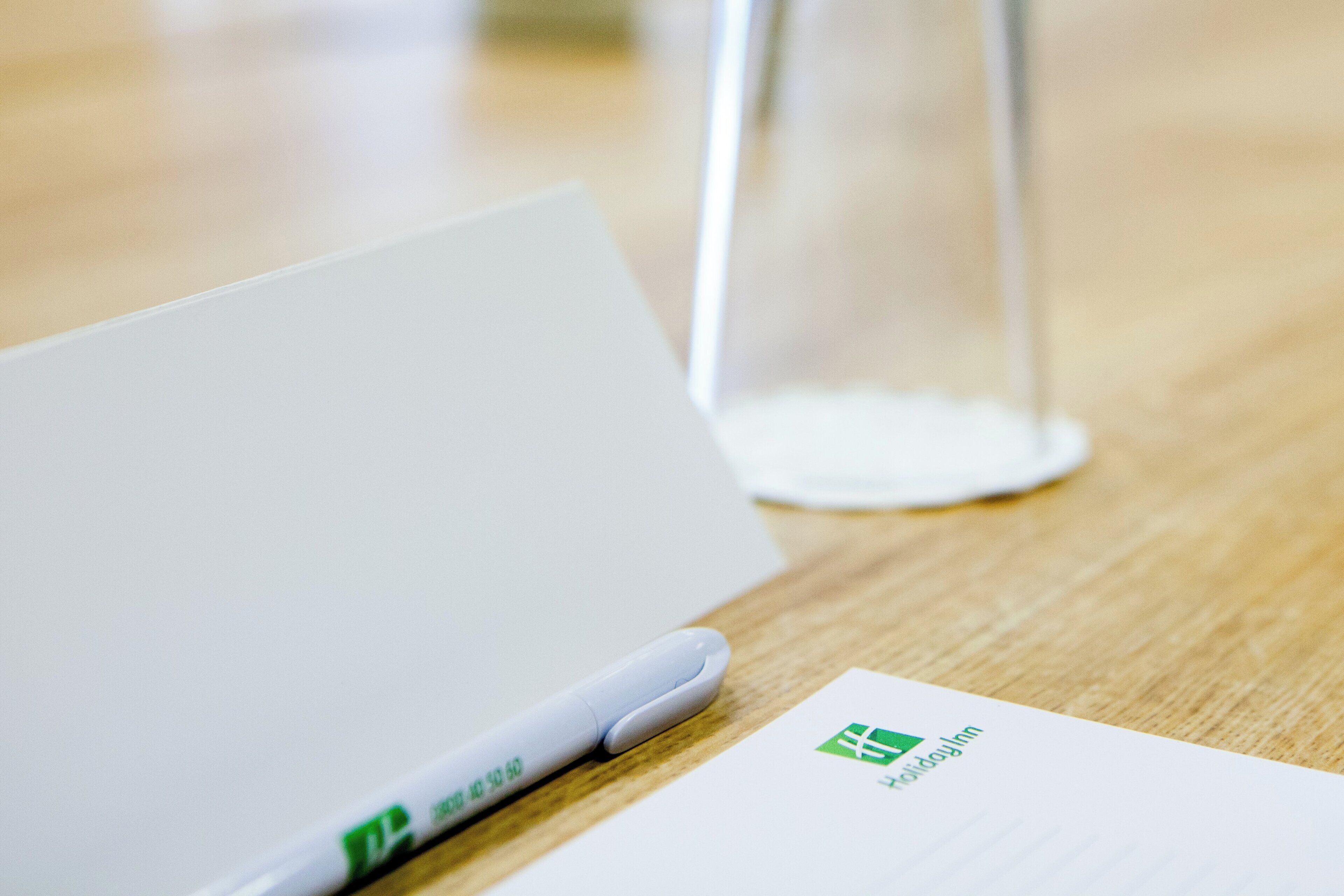 A white notepad with a pen on a wooden table, a clear glass, and a piece of paper with a logo.