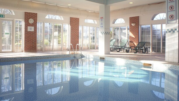 A pool area with clear glass walls, a brick wall, and lounge chairs. - Holiday Inn Corby Kettering Hotel & Spa (Corby)