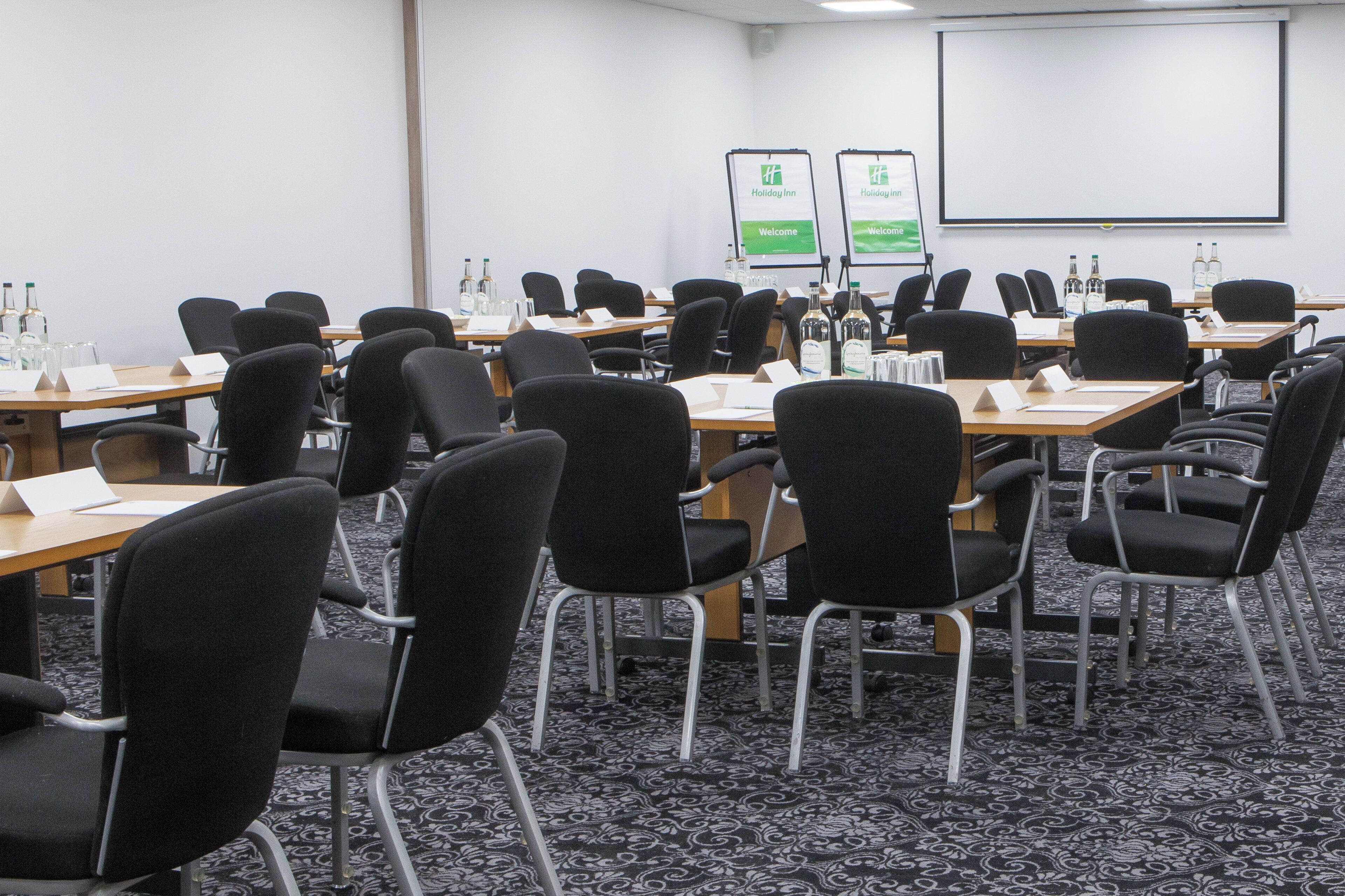 A conference room with rows of black chairs, tables with nameplates, and a projector screen.
