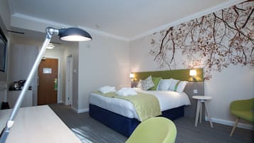 A hotel room with a bed, a desk, a chair, a lamp, and a wall with a tree mural.
