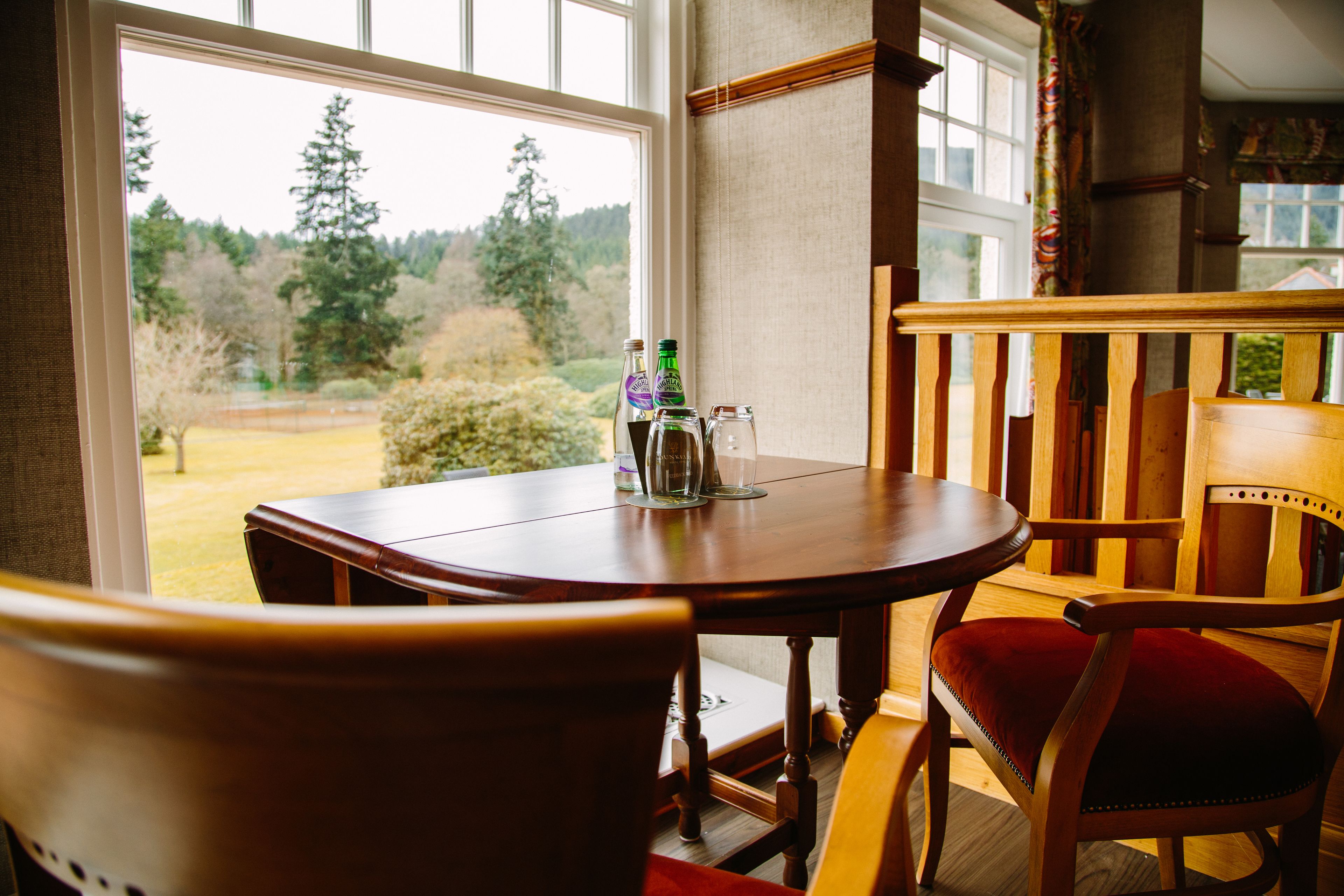 Una mesa de madera con sillas, una ventana que da a árboles y un césped, y una botella de cerveza sobre la mesa.