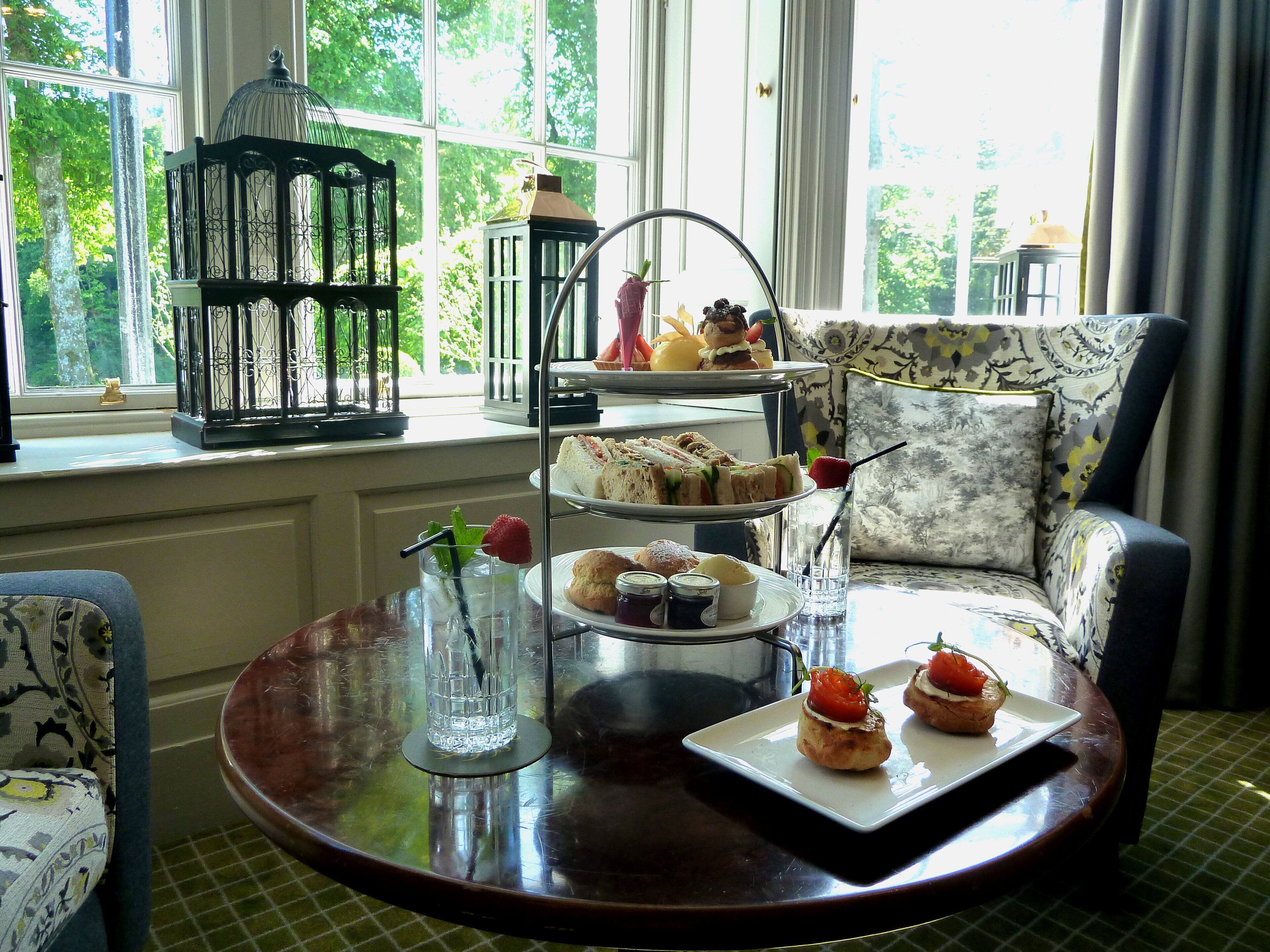 A dining area with a three-tiered dessert stand, a table with a glass of iced tea and a plate of pastries, and a patterned armchair.