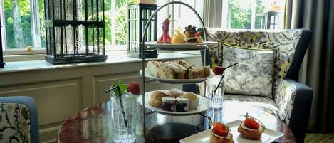 A dining area with a three-tiered dessert stand, a table with a glass of iced tea and a plate of pastries, and a patterned armchair.