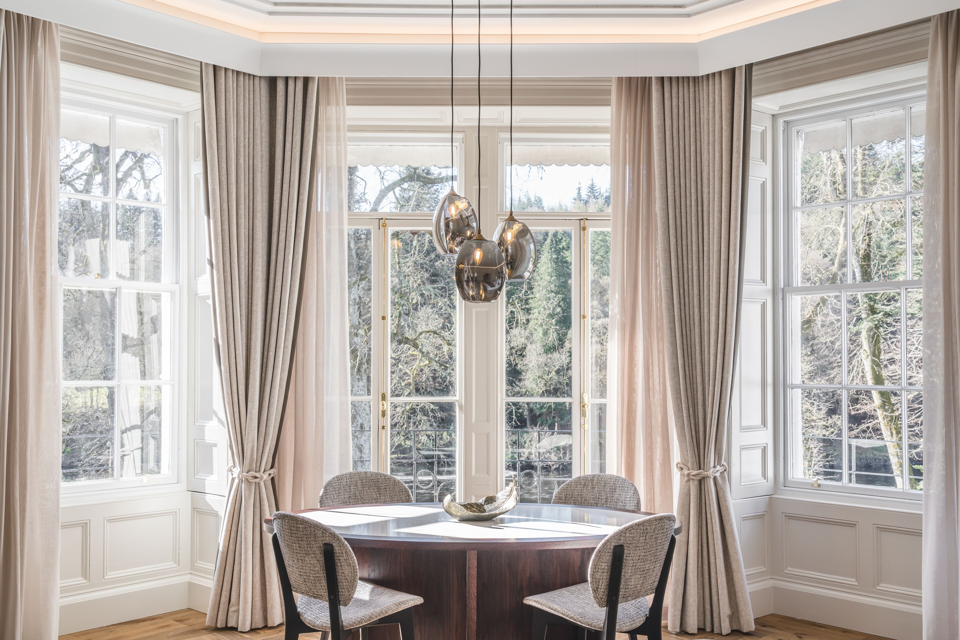 Uma área de refeições com uma mesa redonda, quatro cadeiras, um candelabro e grandes janelas com cortinas.