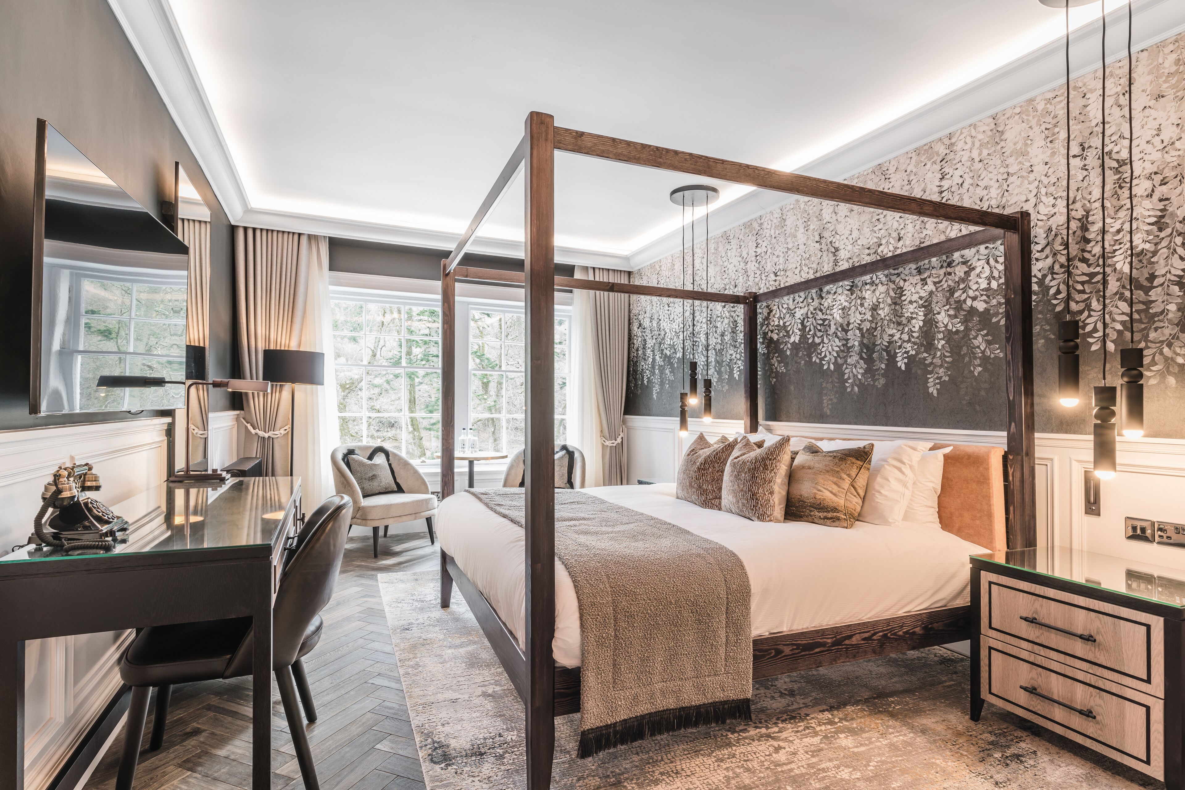 A four-poster bed with a wooden frame, a desk with a chair, a bedside table, and a large window with curtains.