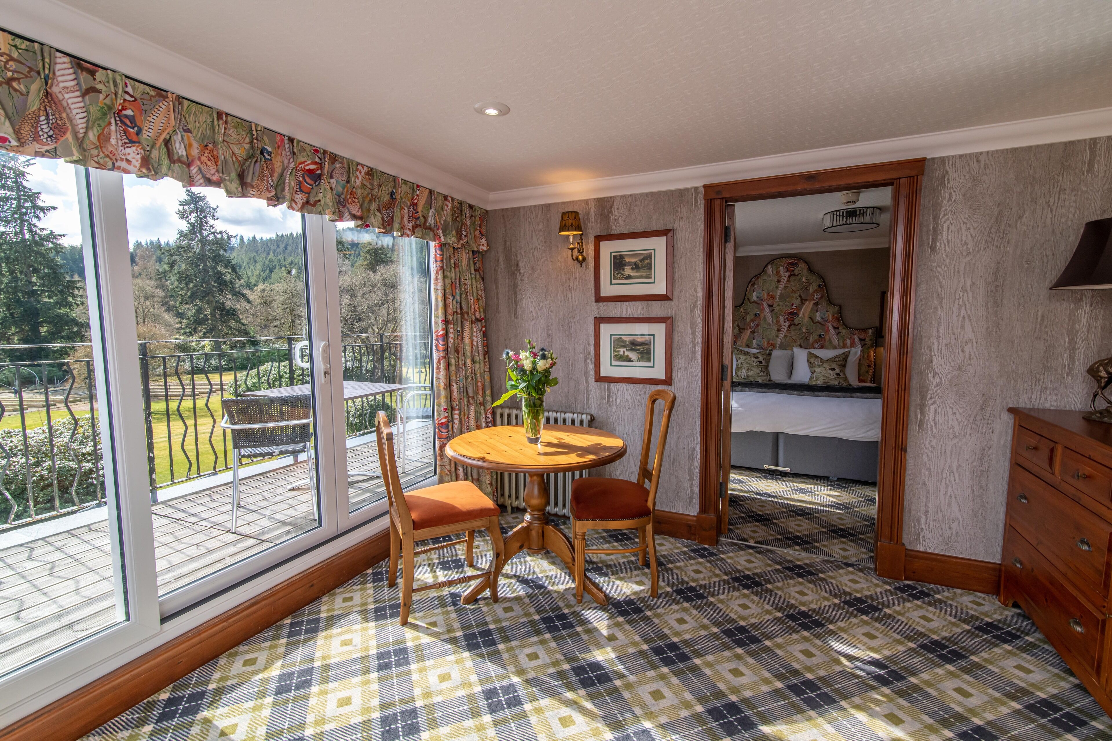 A room with a checkered floor, a round wooden table with chairs, a bed with a floral headboard, and a view of a garden through large windows.