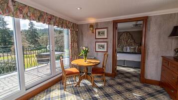 A room with a checkered floor, a round wooden table with chairs, a bed with a floral headboard, and a view of a garden through large windows.