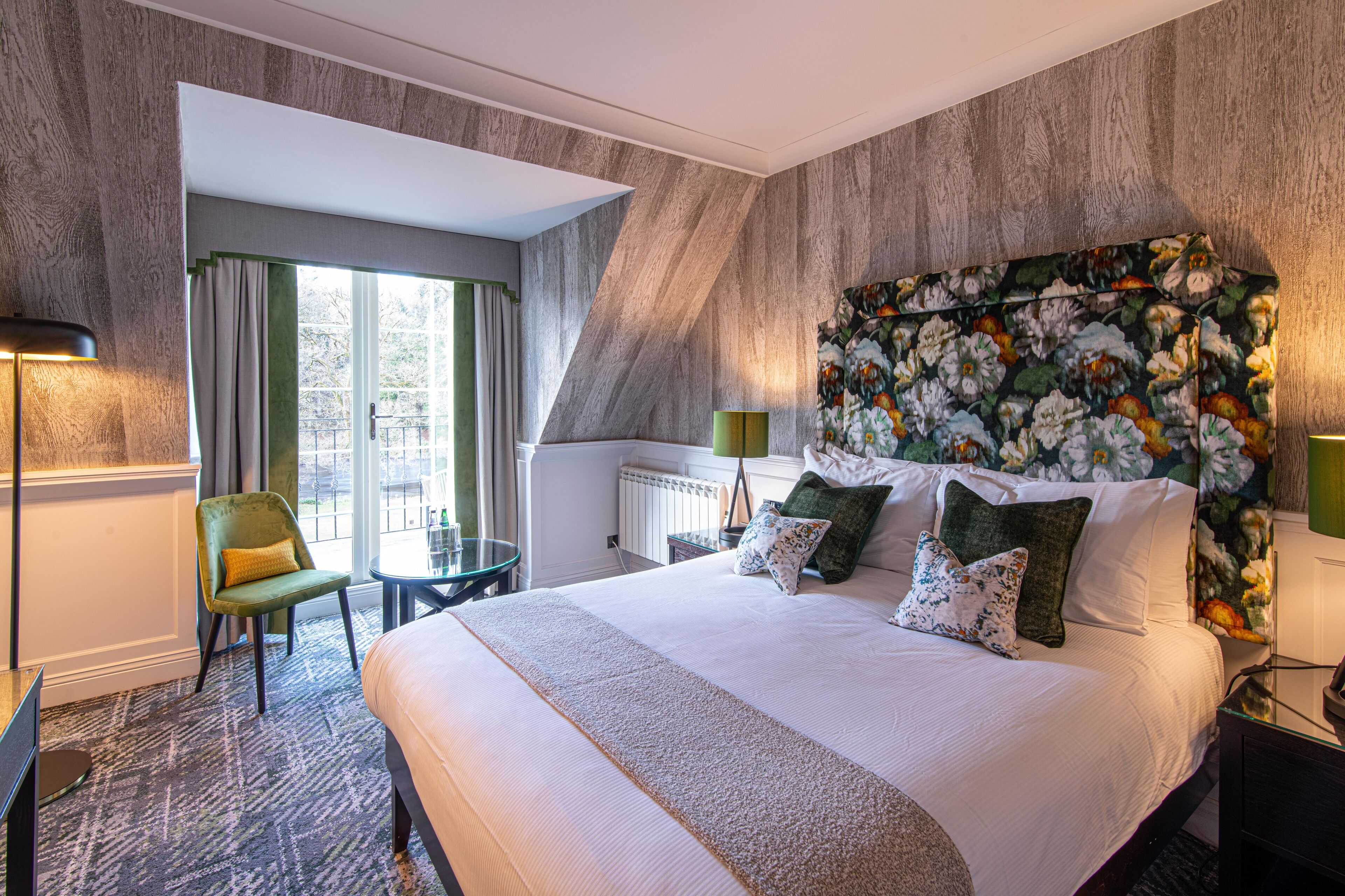 A hotel room with a large bed, a green chair, a small table, and a view of the outdoors.