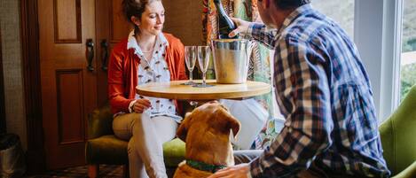 A room with a wooden door, a green armchair, a wooden table with a bucket and glasses, and a dog.