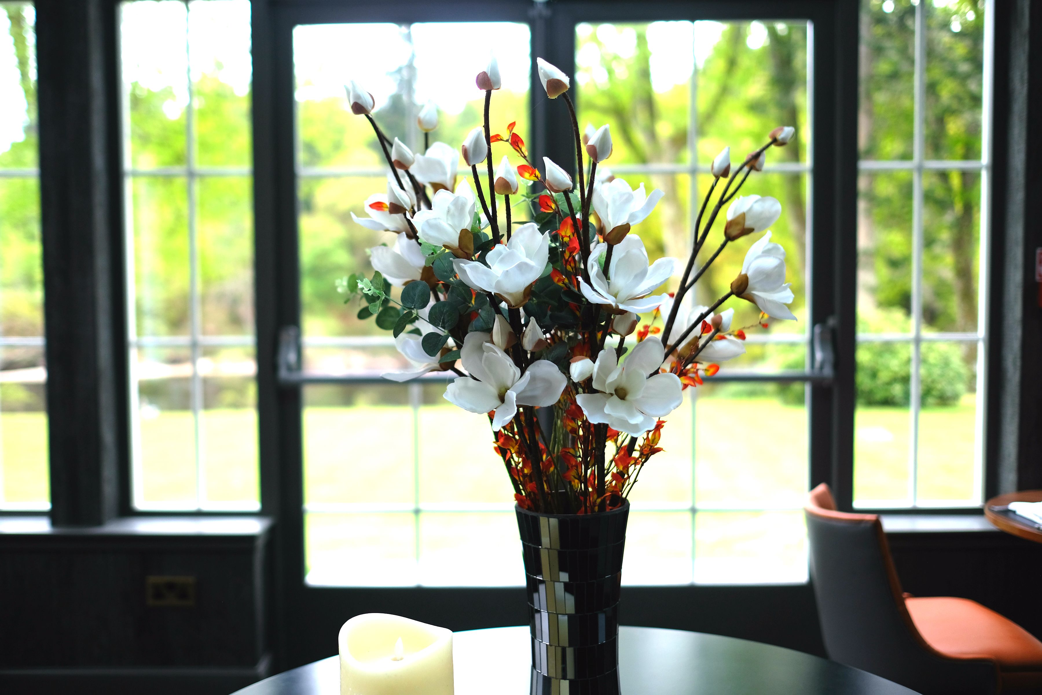 Un jarrón con flores blancas y una vela encendida sobre una mesa junto a una ventana.