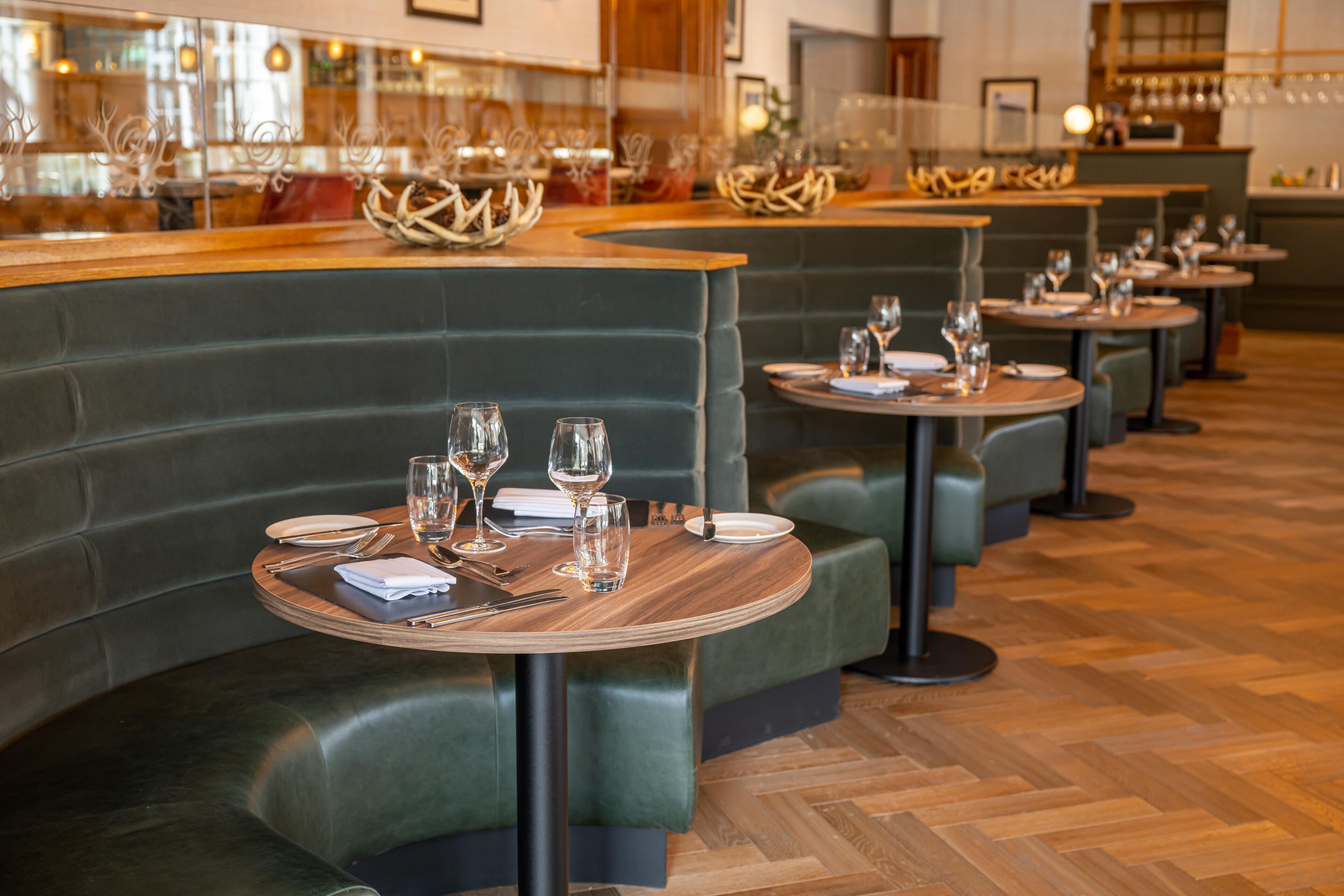 Ein Restaurant mit grünen Sitzbänken und runden Tischen, an jedem Tisch Weingläser und Besteck.