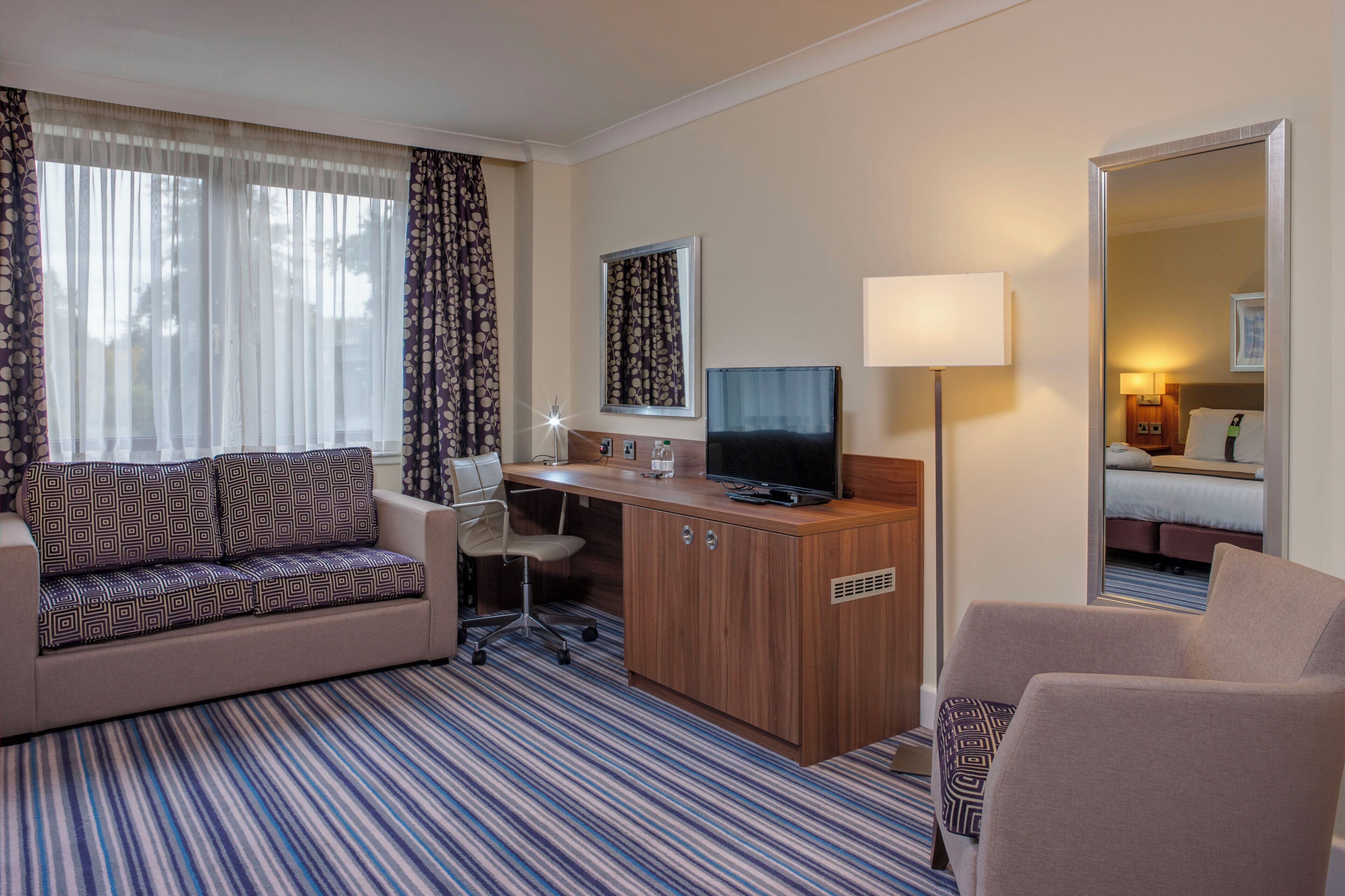 A hotel room with a sofa, a desk with a computer, a chair, a bed, and a mirror.