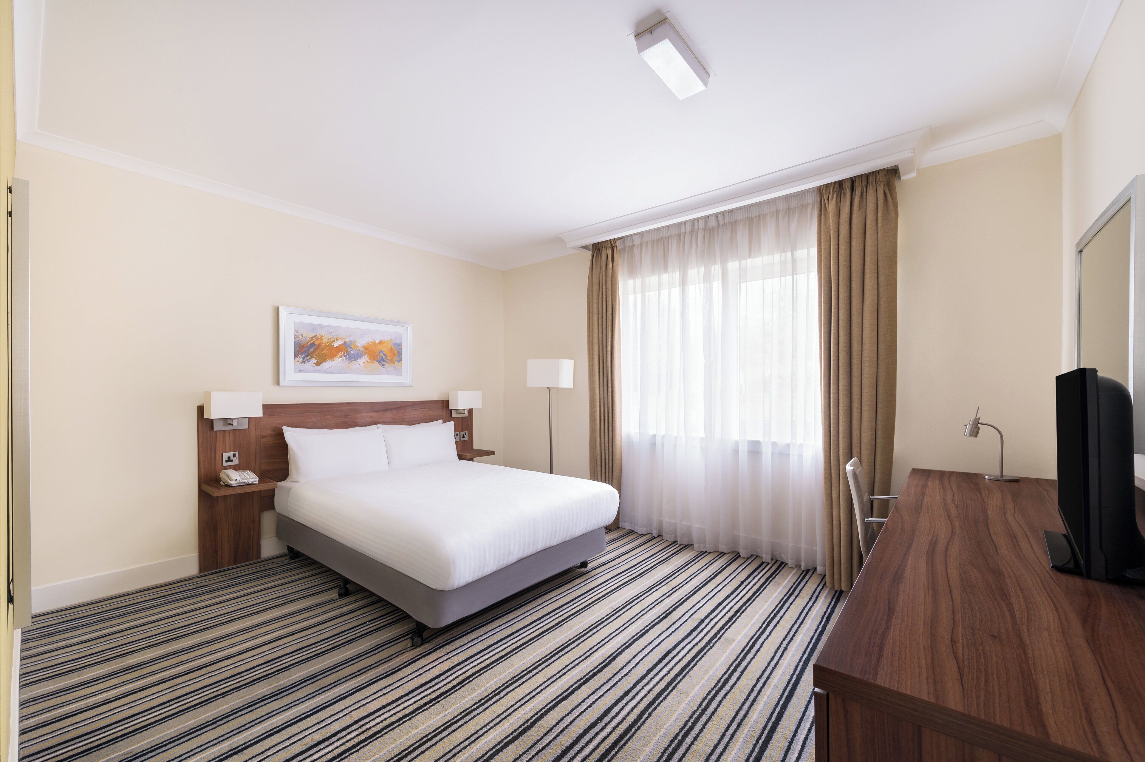 Habitación de hotel con cama, escritorio, televisor, ventana con cortinas y un cuadro en la pared.