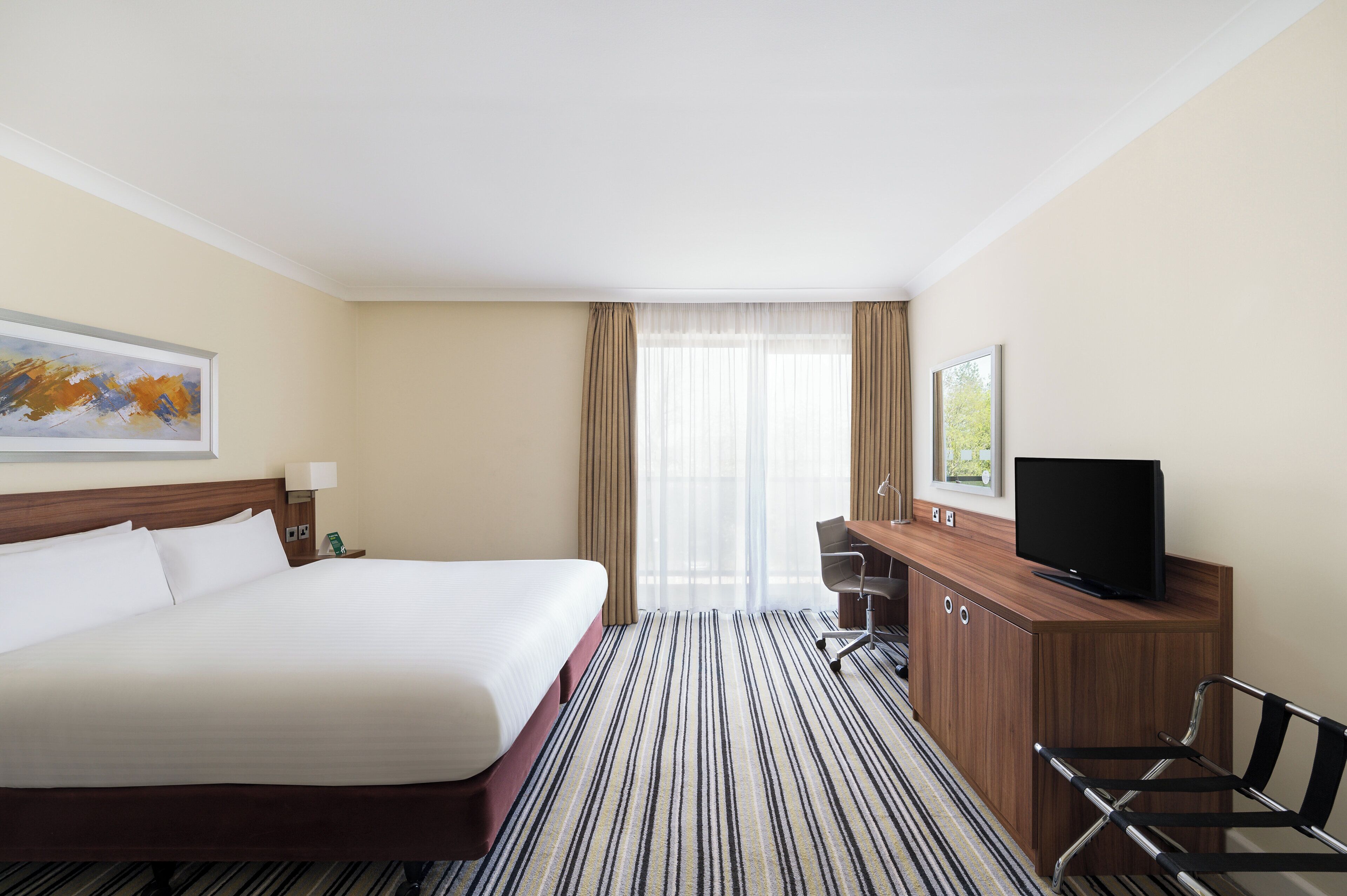 A hotel room with a large bed, a desk with a flat-screen TV, a chair, and a window with sheer curtains.