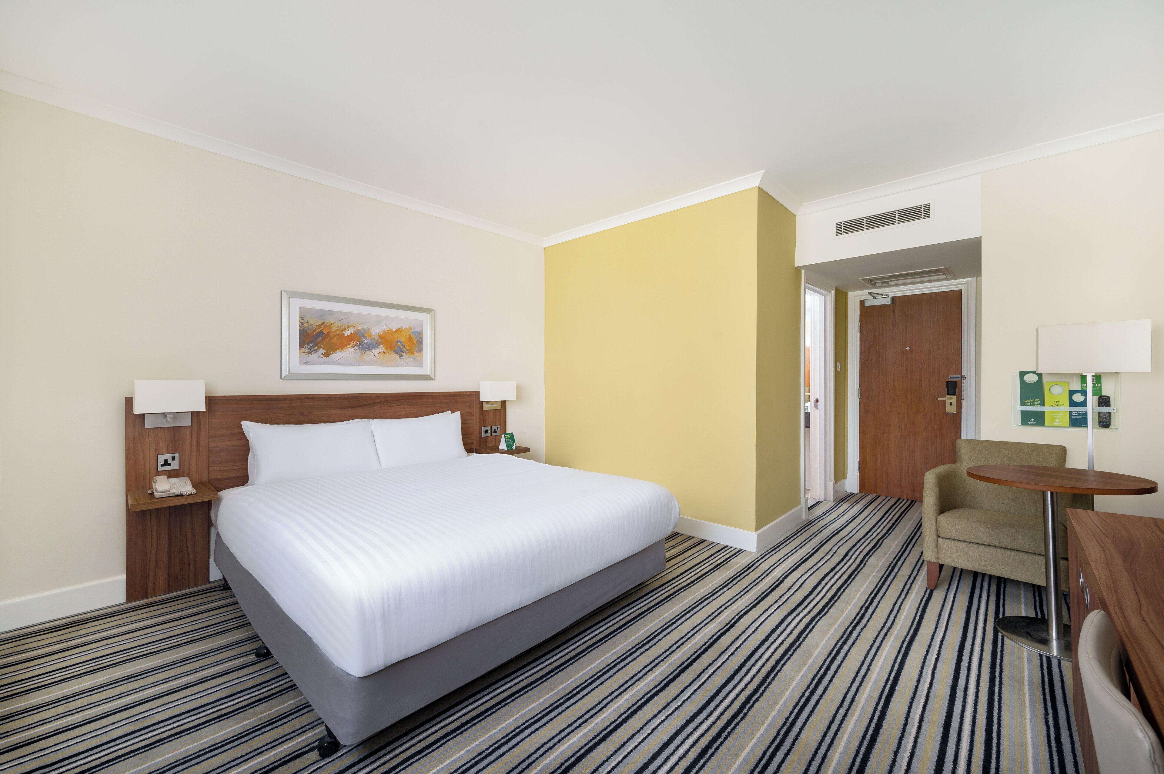 Ein Hotelzimmer mit einem großen Bett, einem kleinen Schreibtisch, einem Stuhl, einem Bild an der Wand und einer Tür zu einem weiteren Raum.