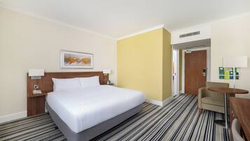 Ein Hotelzimmer mit einem großen Bett, einem kleinen Schreibtisch, einem Stuhl, einem Bild an der Wand und einer Tür zu einem weiteren Raum.