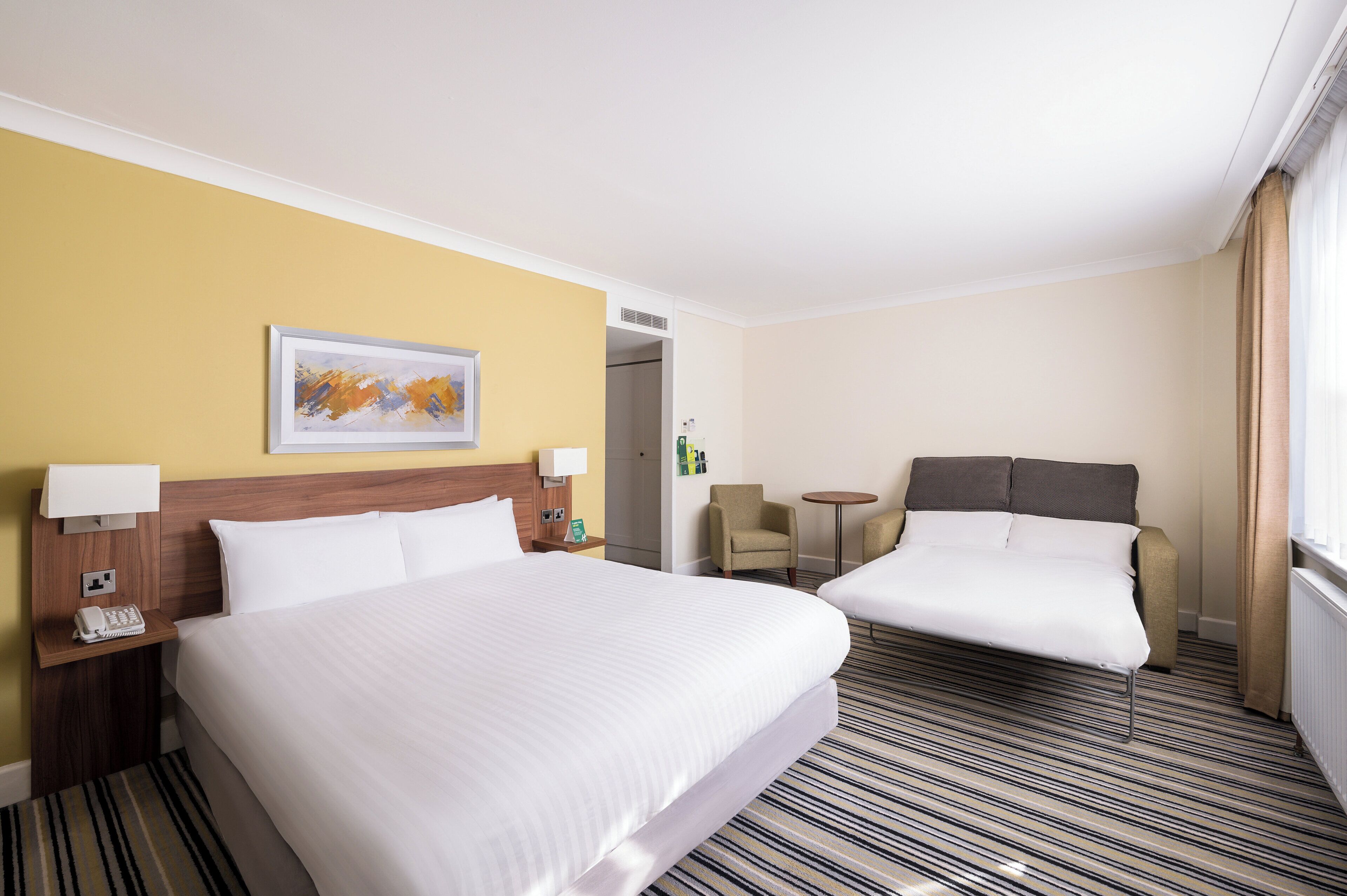 A hotel room with two beds, a small table, a chair, and a painting on the wall.