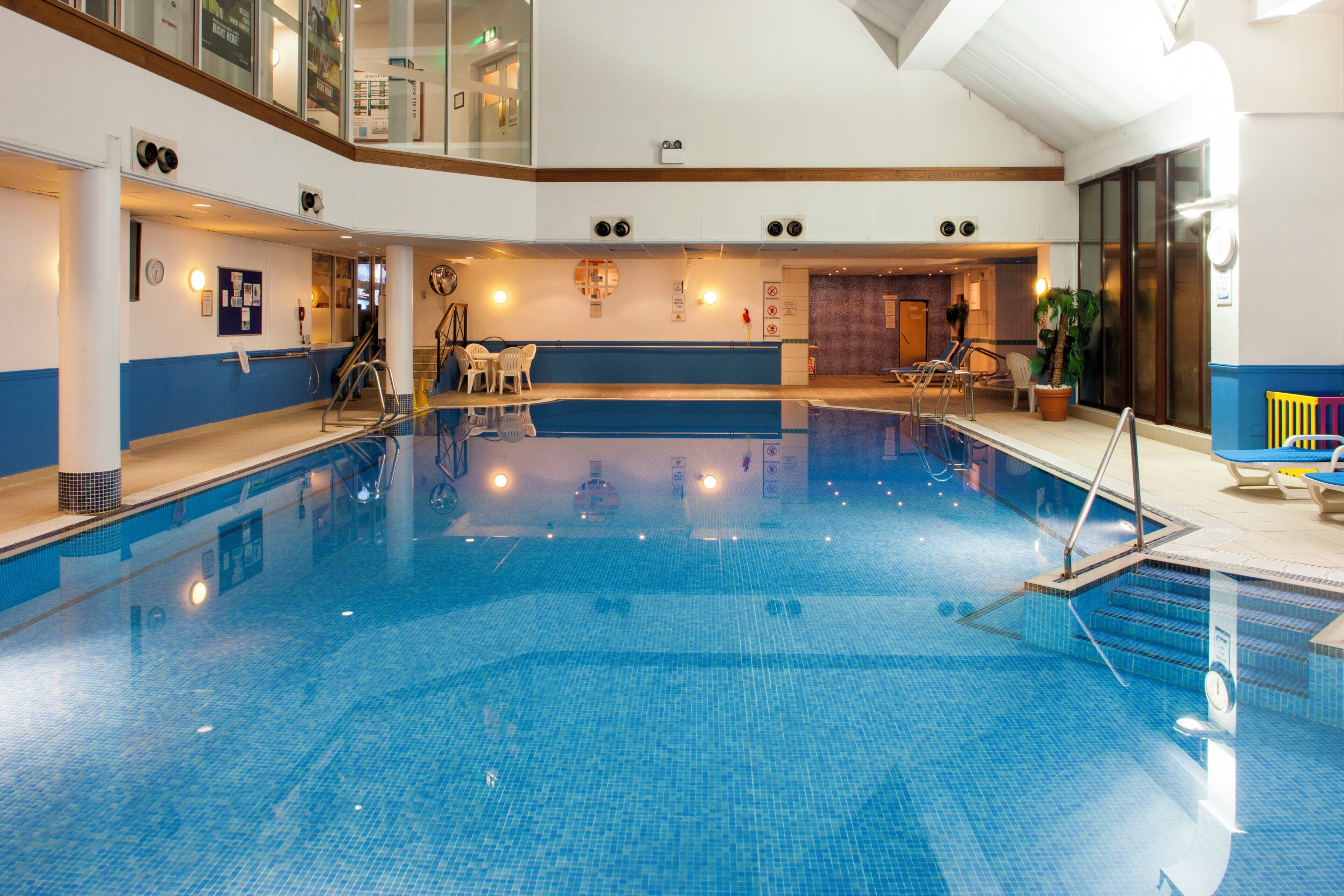 An indoor swimming pool with a clear view of the pool area, including the pool's edge, a seating area with chairs, and a staircase leading to an upper level.