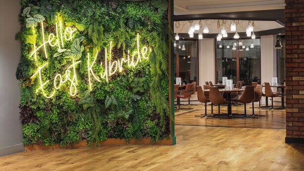 A modern interior with a neon sign reading "Hello, I love Kilbride" against a lush green wall, wooden flooring, and a dining area with chairs and tables. - Holiday Inn Glasgow East Kilbride by IHG (Glasgow)