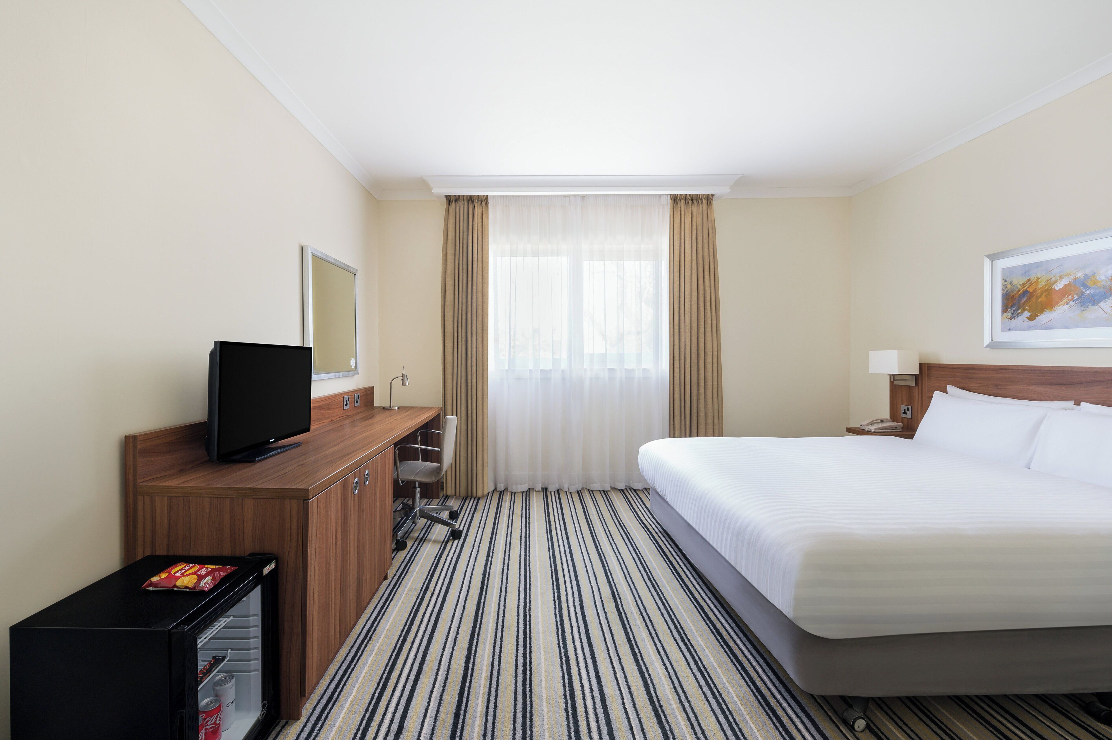 Habitación de hotel con cama, escritorio con televisor, mini refrigerador y ventana con cortinas.
