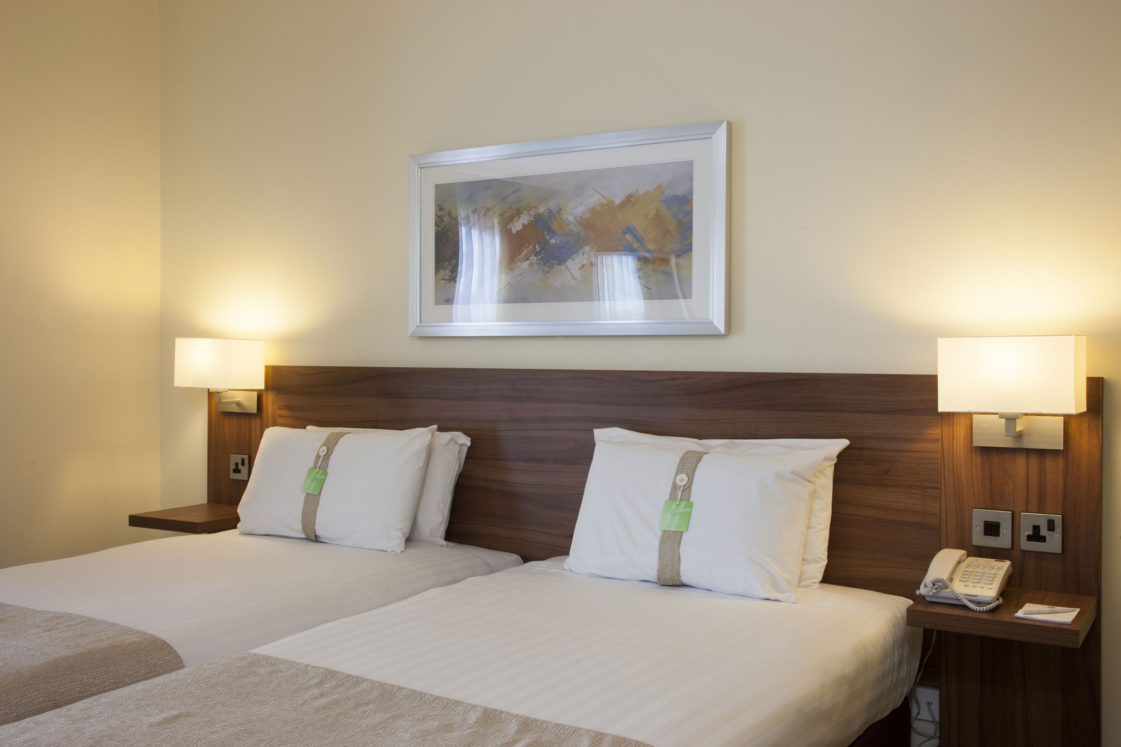 A hotel room with two beds, a wooden headboard, bedside lamps, a painting on the wall, and a telephone on the nightstand.