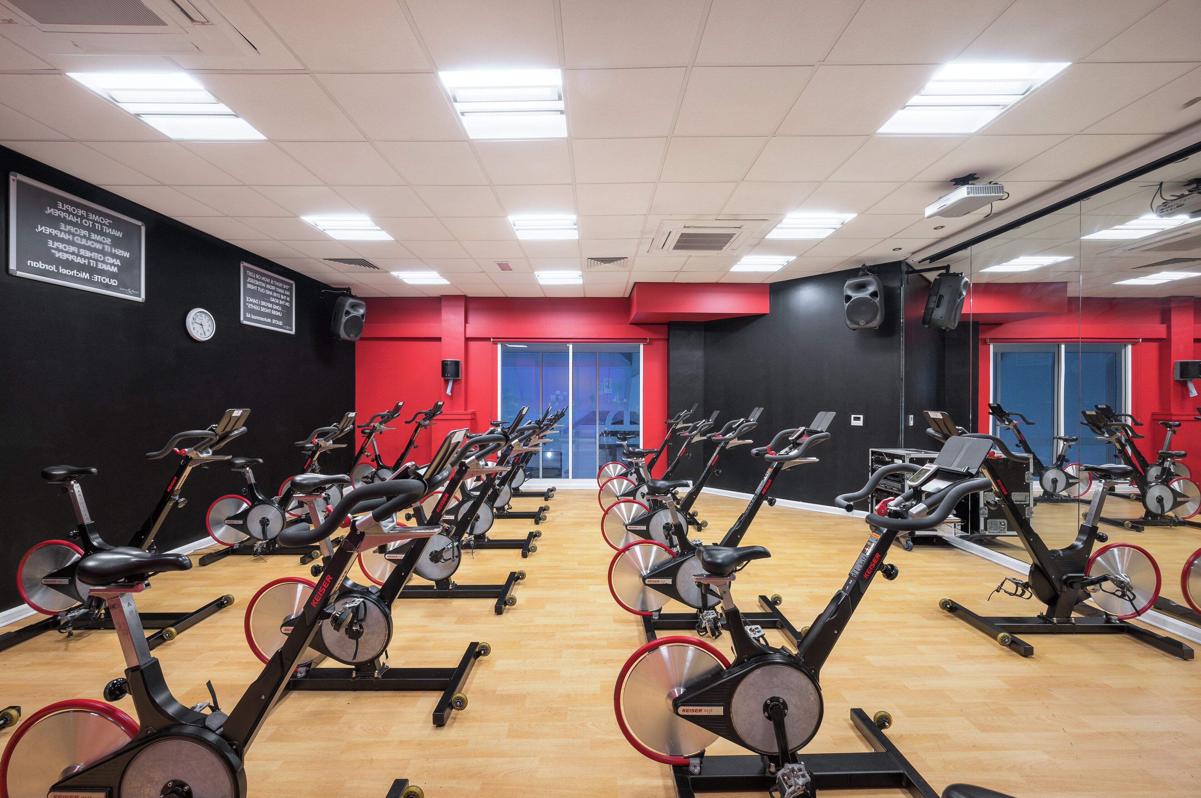 Ein Fitnessraum mit mehreren in Reihen angeordneten Ergometer, einer Uhr an der Wand und einem Farbkonzept in Rot und Schwarz.
