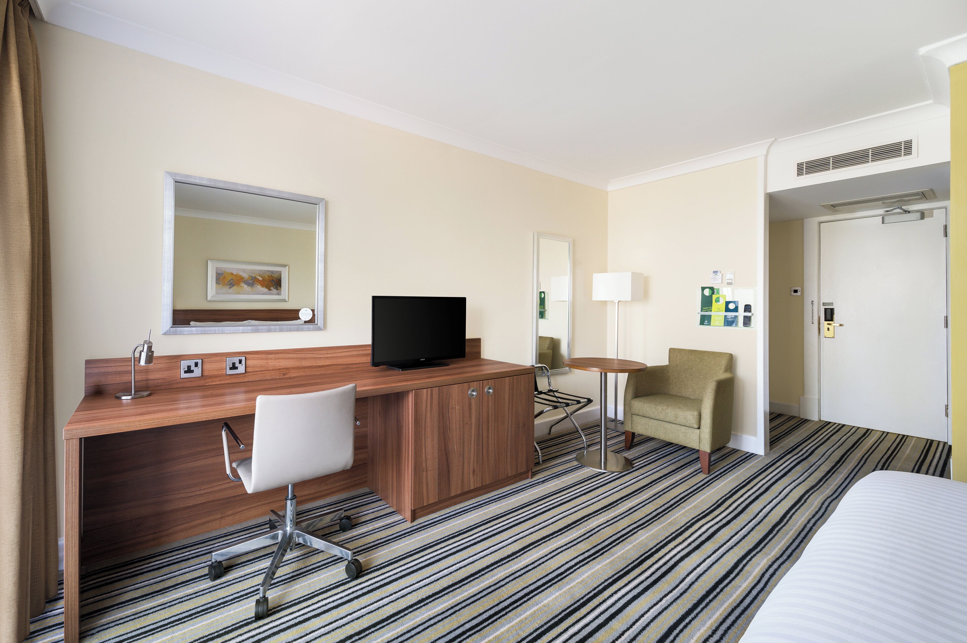 A hotel room with a wooden desk, a television, a chair, a bed, and a striped carpet.