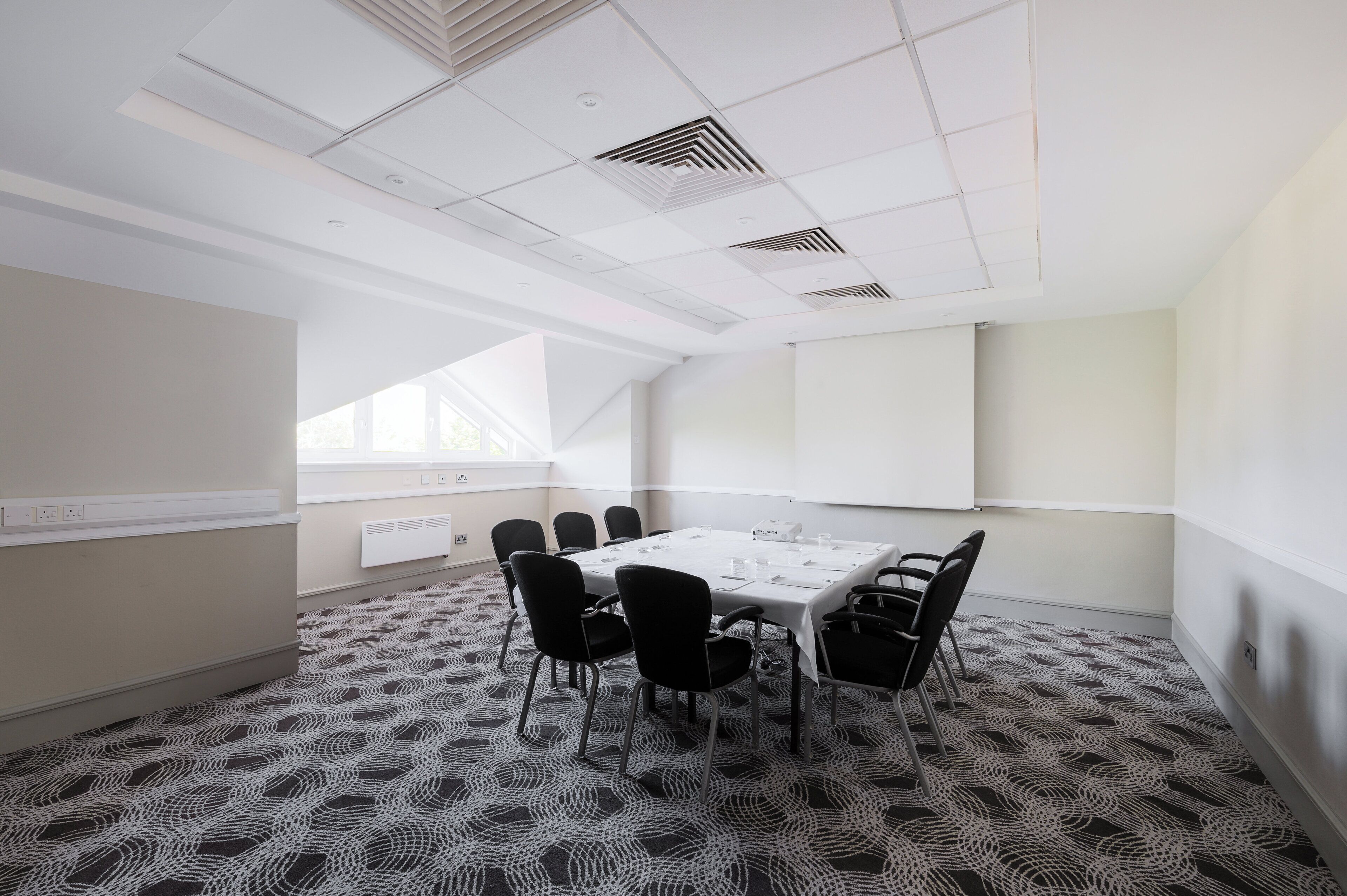 A conference room with a large table, black chairs, patterned carpet, white walls, and a window with blinds.