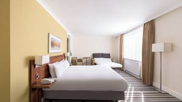 Ein Hotelzimmer mit einem großen Bett, Nachttischen, einem kleinen Schreibtisch, einem Sessel und einem Fenster mit Vorhängen.